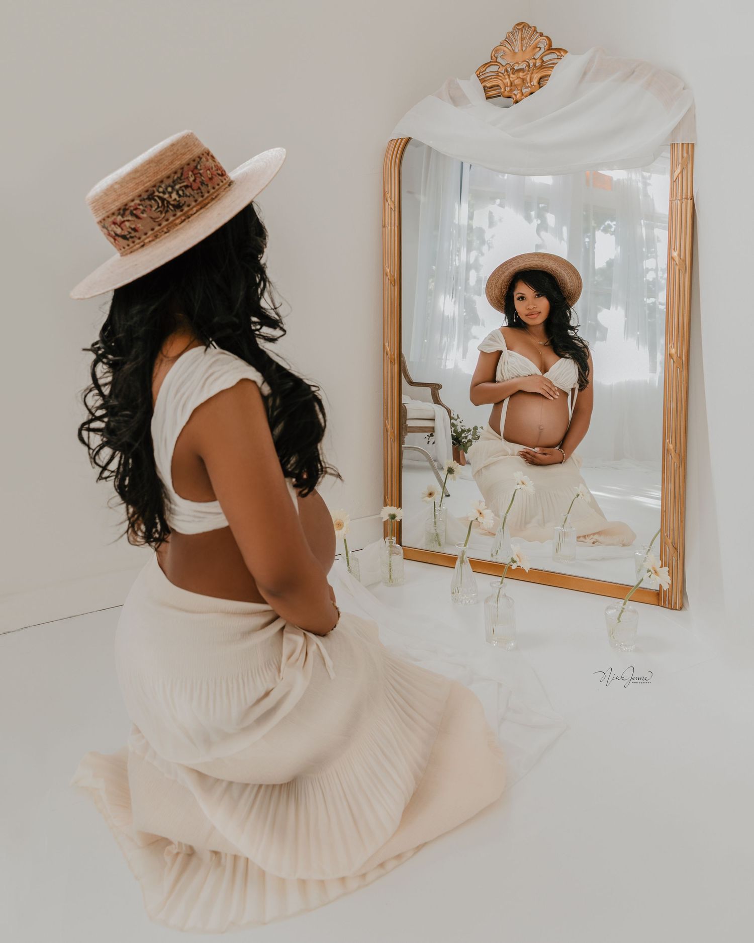 Elegant maternity photoshoot featuring white flowing dress and straw hat against light backdrop.