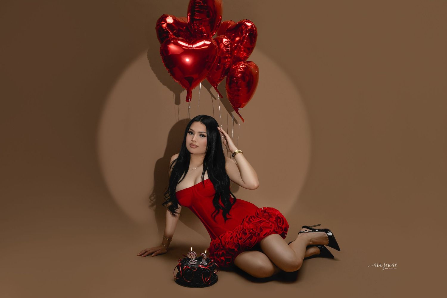 Woman in black lingerie poses with red heart-shaped balloons against a brown backdrop.