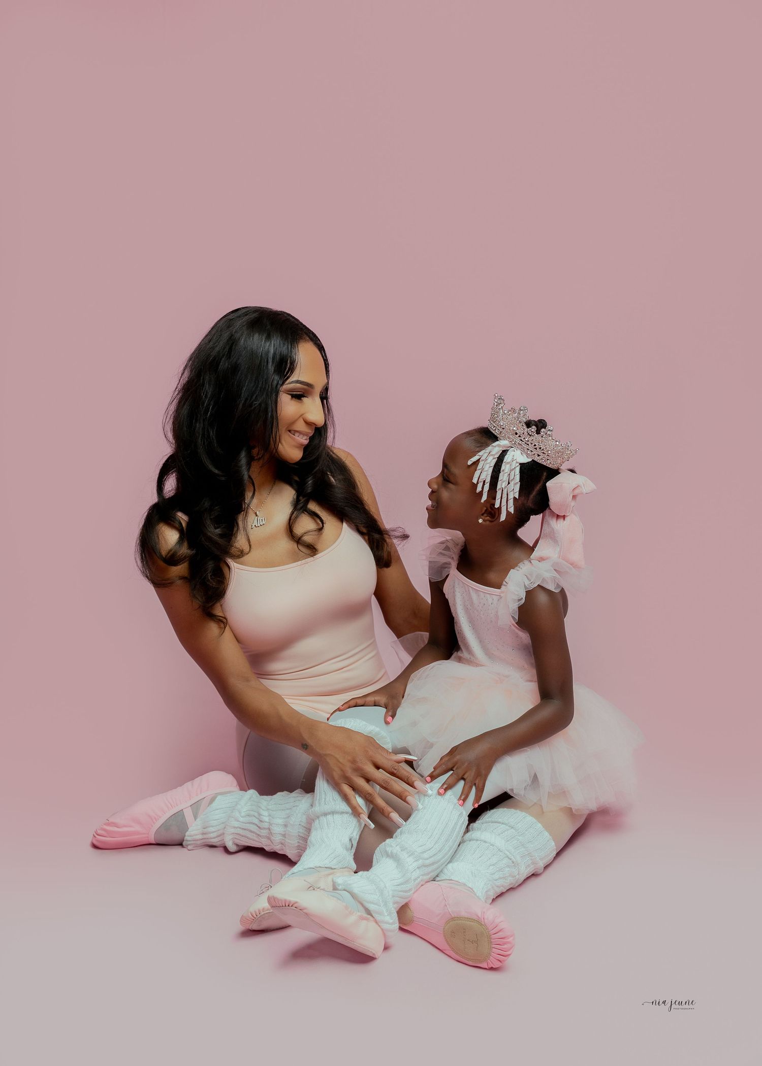 A woman and young girl in pink ballet outfits sit together smiling on a pink studio background.