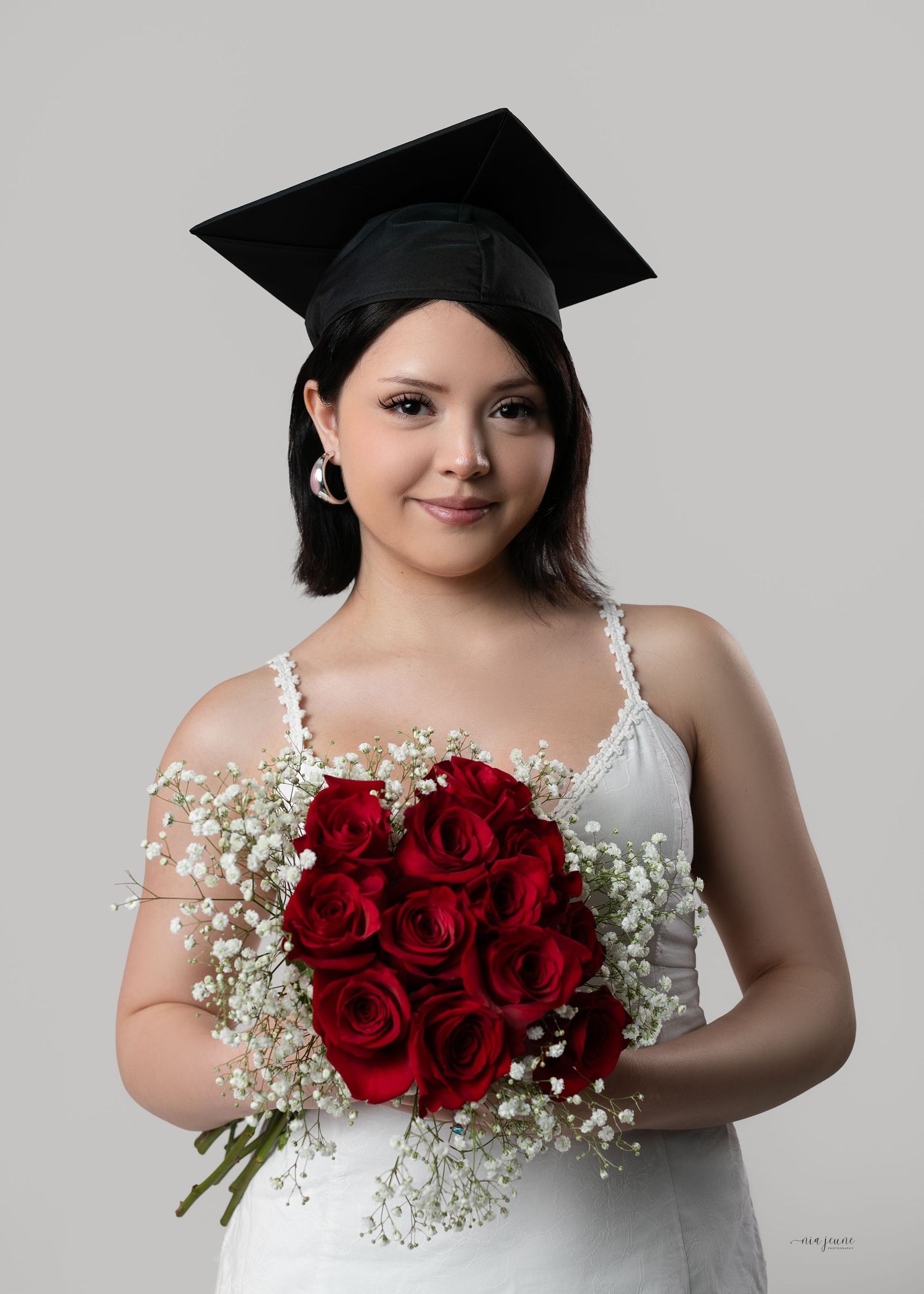 Young woman in graduation cap holding heart-shaped red rose bouquet with baby's breath, wearing white dress.