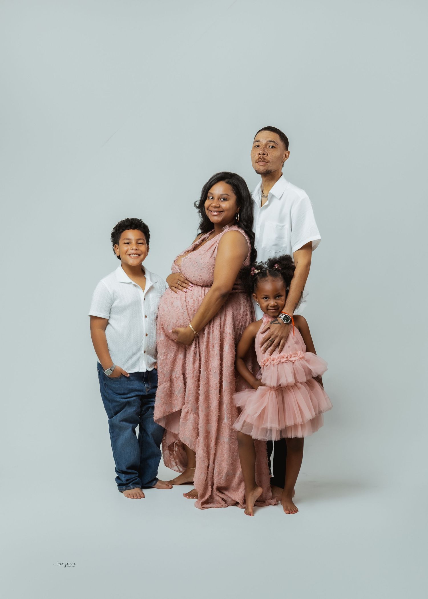 Pregnant woman in pink lace gown poses with family including two children and partner in a studio maternity photo.