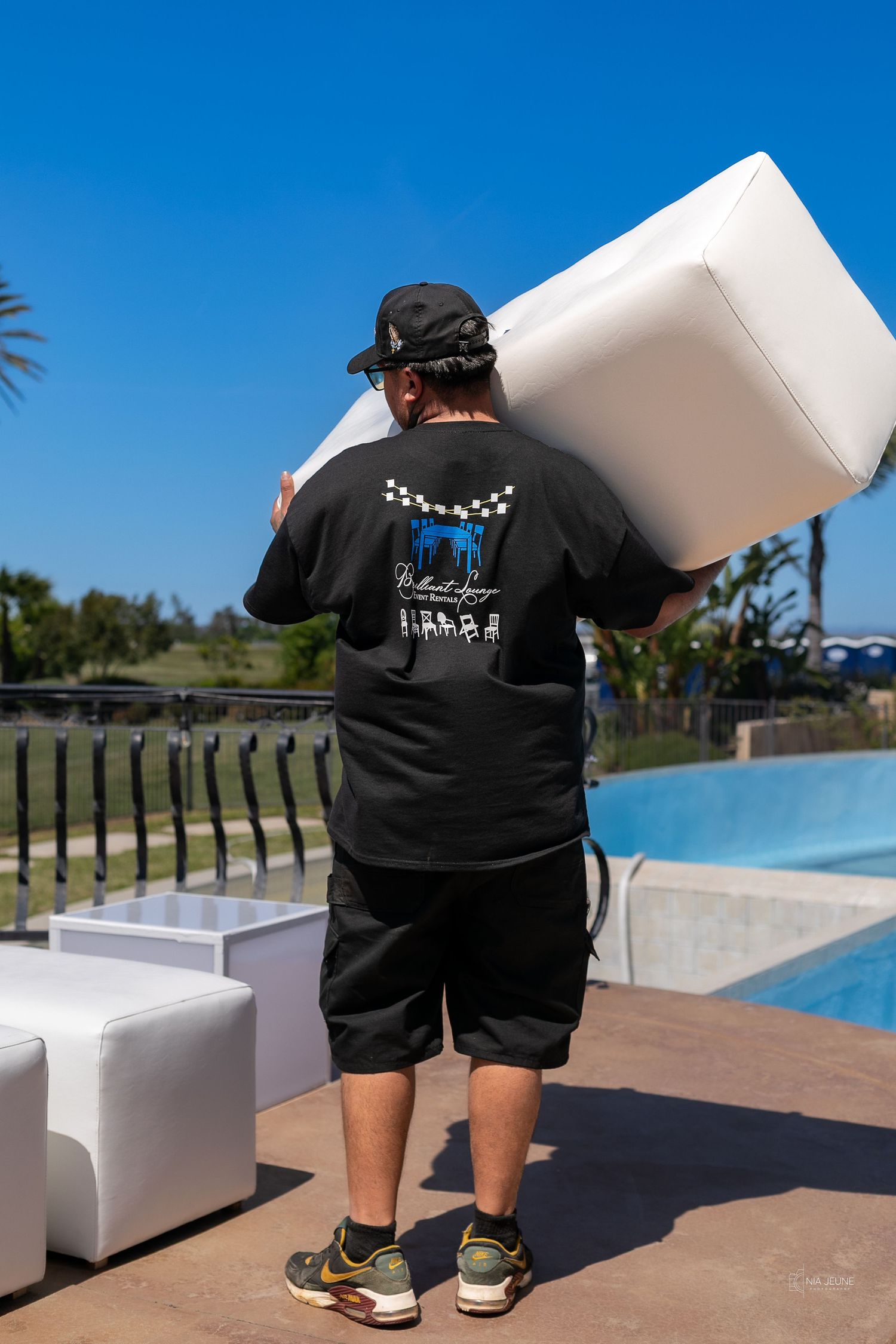 Man in black t-shirt carrying white cube ottoman near a pool on a sunny day.