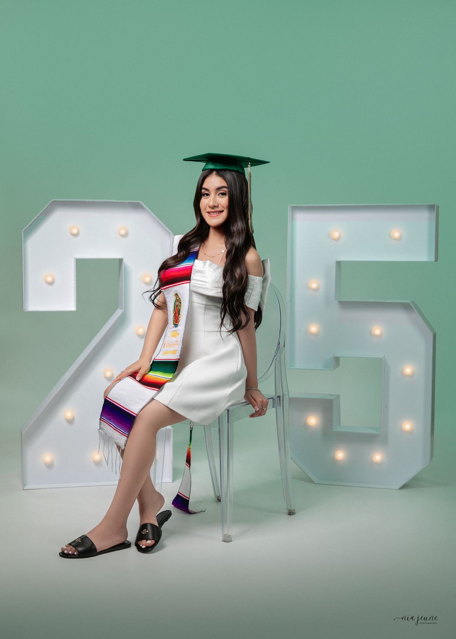 Graduate in white dress and cap poses by illuminated '25' numbers against mint green backdrop.