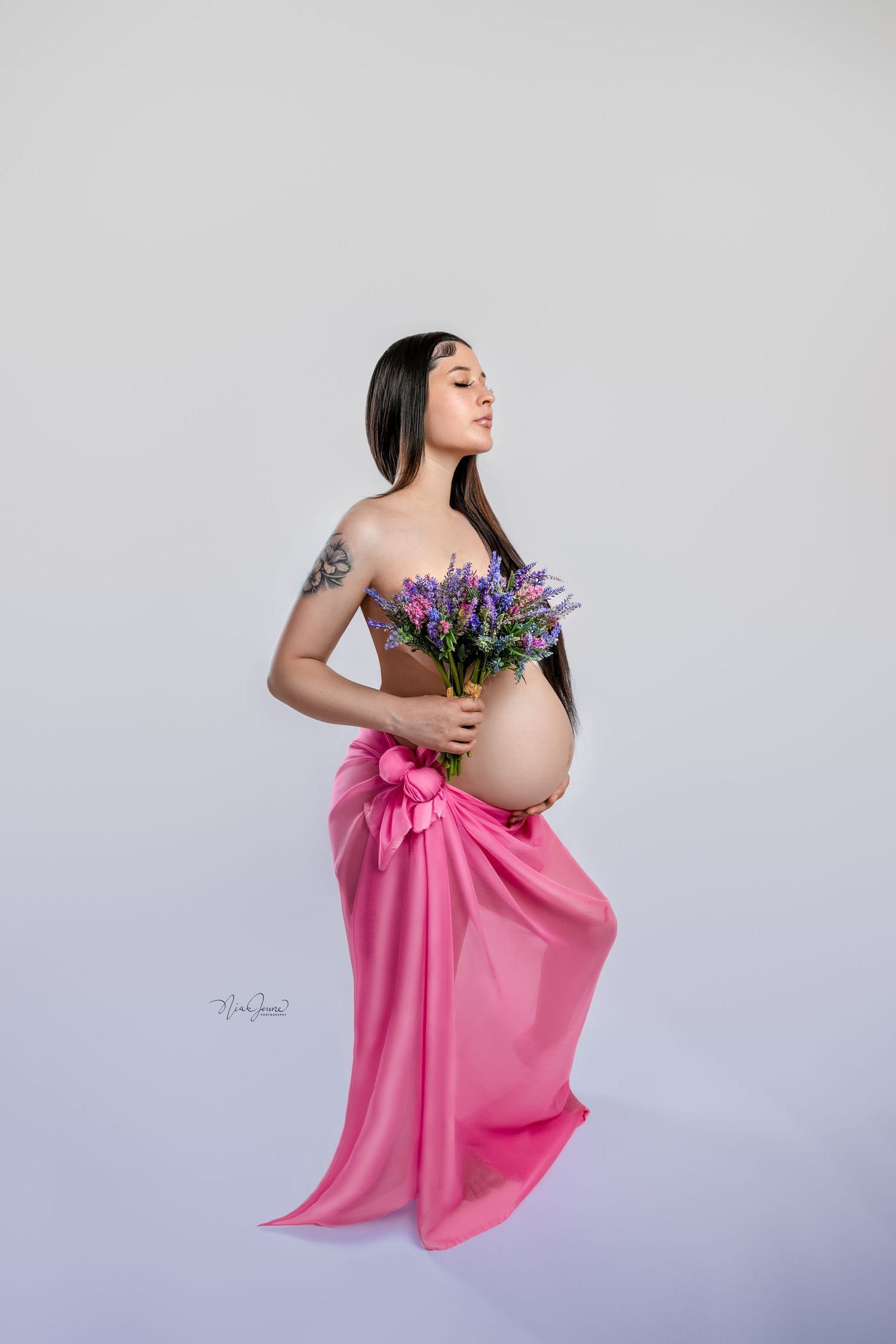 Expectant mother in pink draping holds purple flowers in elegant maternity photo.