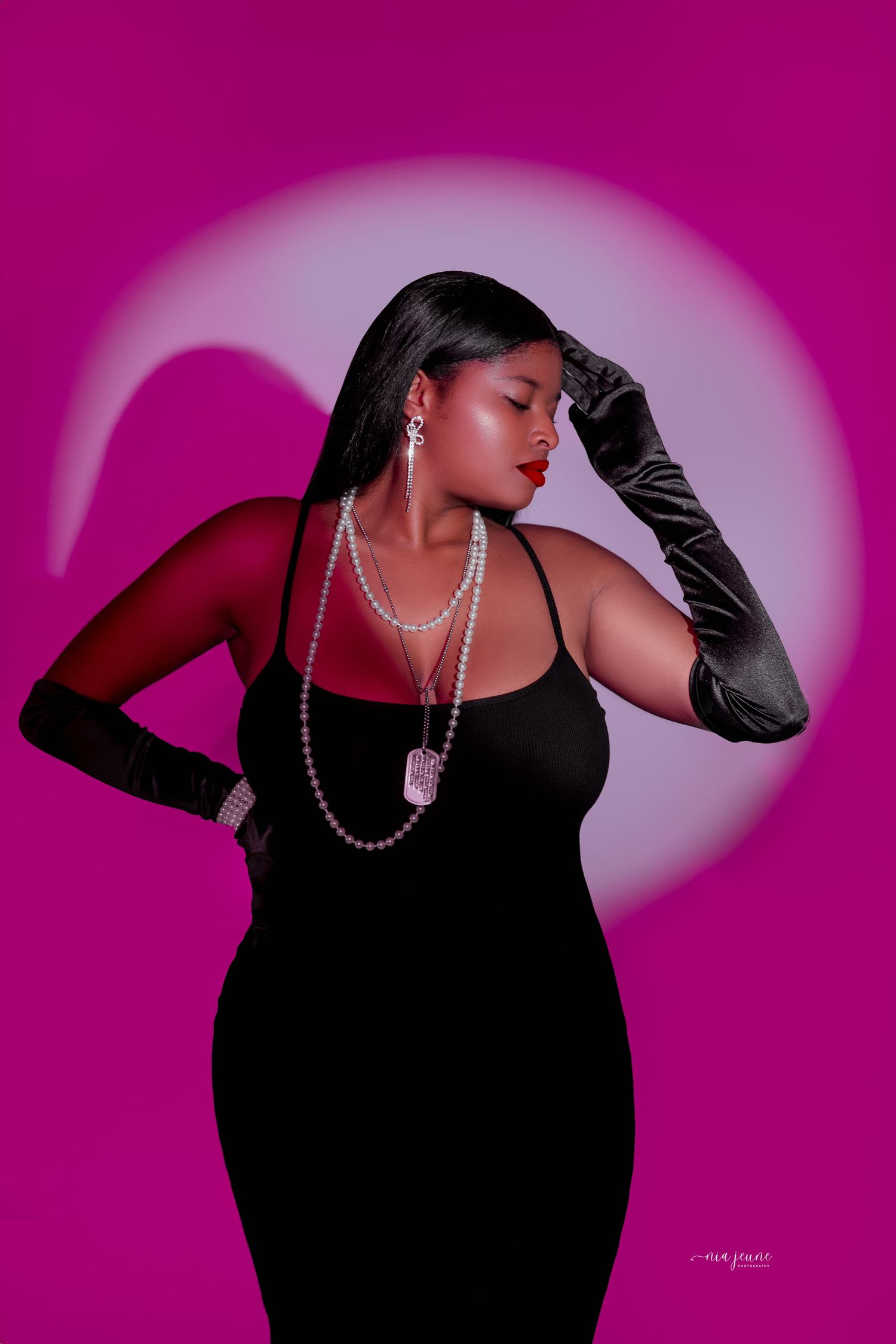 Dramatic fashion portrait with long black dress, pearls, and gloves against magenta background.