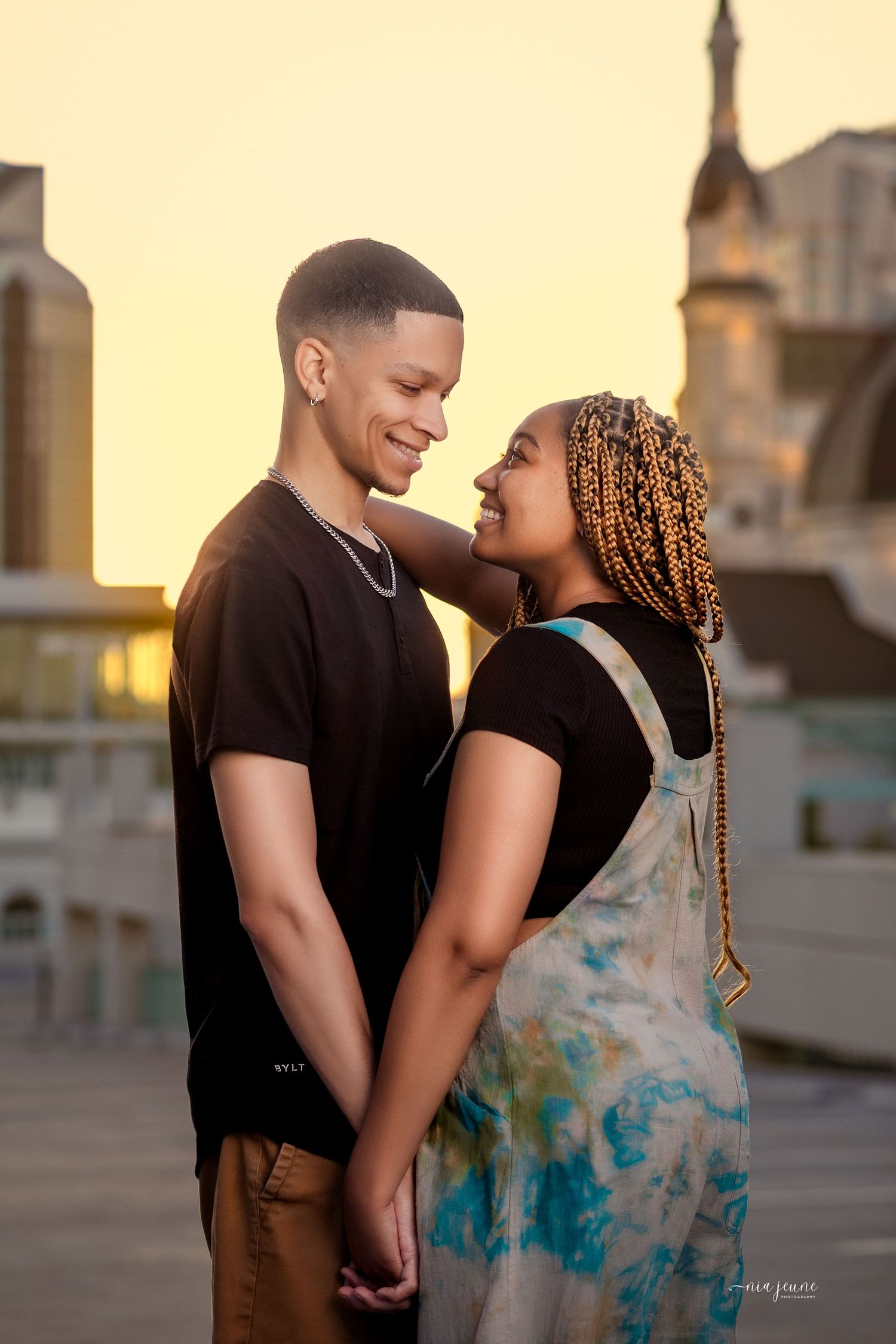 Romantic couple in matching black outfits share tender moments against a sunset urban backdrop.