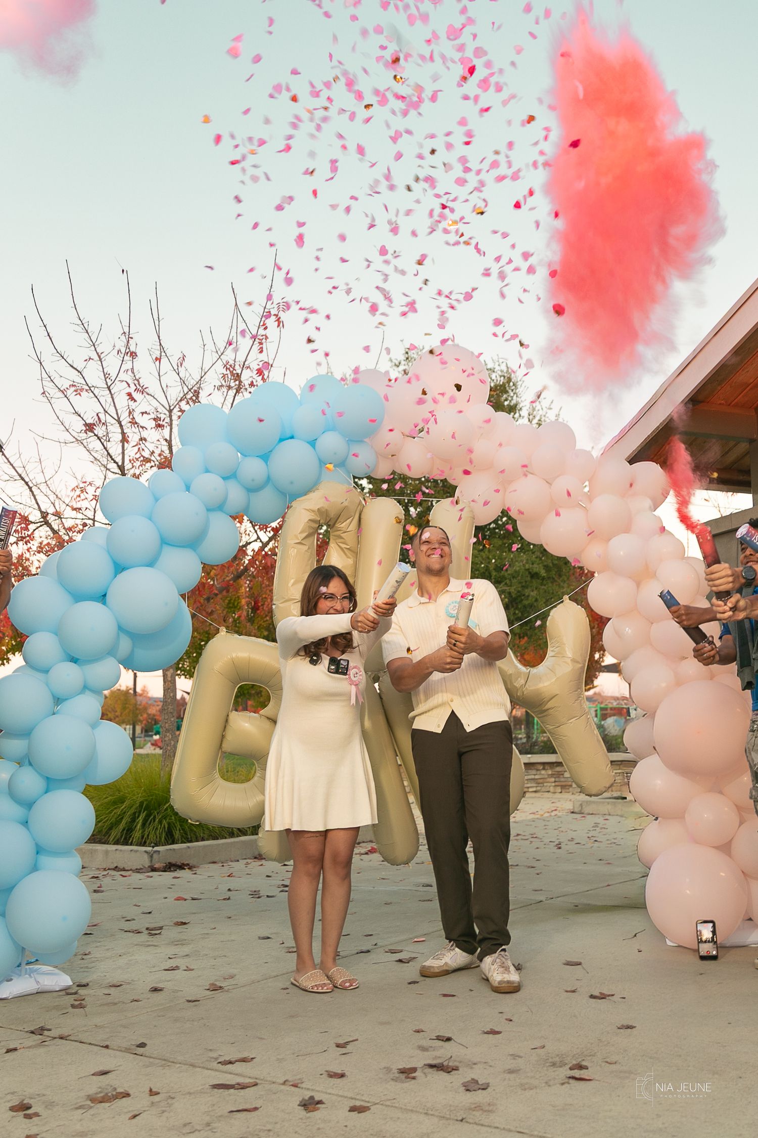Couple celebrates gender reveal with pink and blue balloon arches, confetti, and colorful smoke at outdoor party.