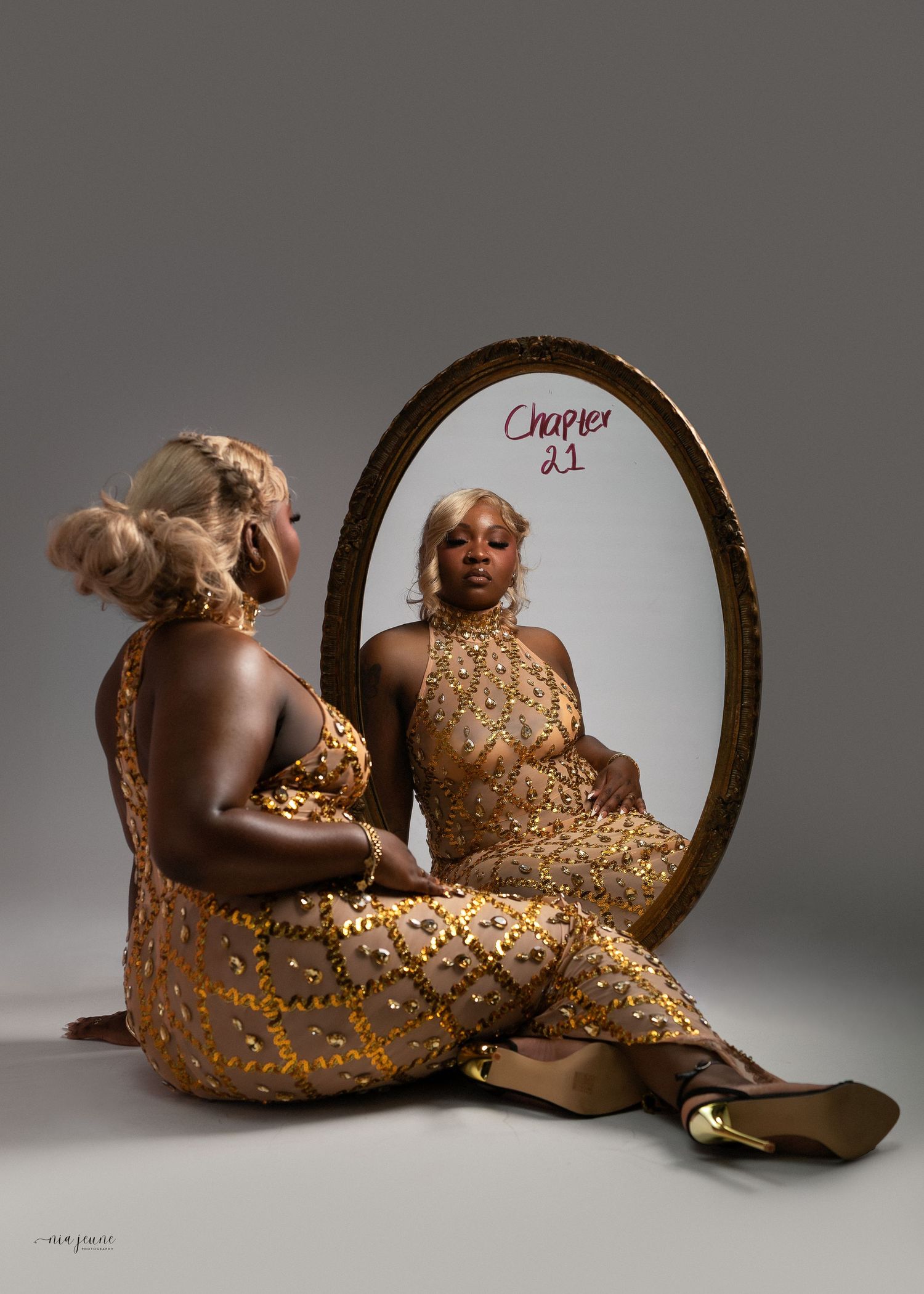 Woman in gold sequin jumpsuit sits before oval mirror labeled Chapter 21, reflecting her image in a studio setting.