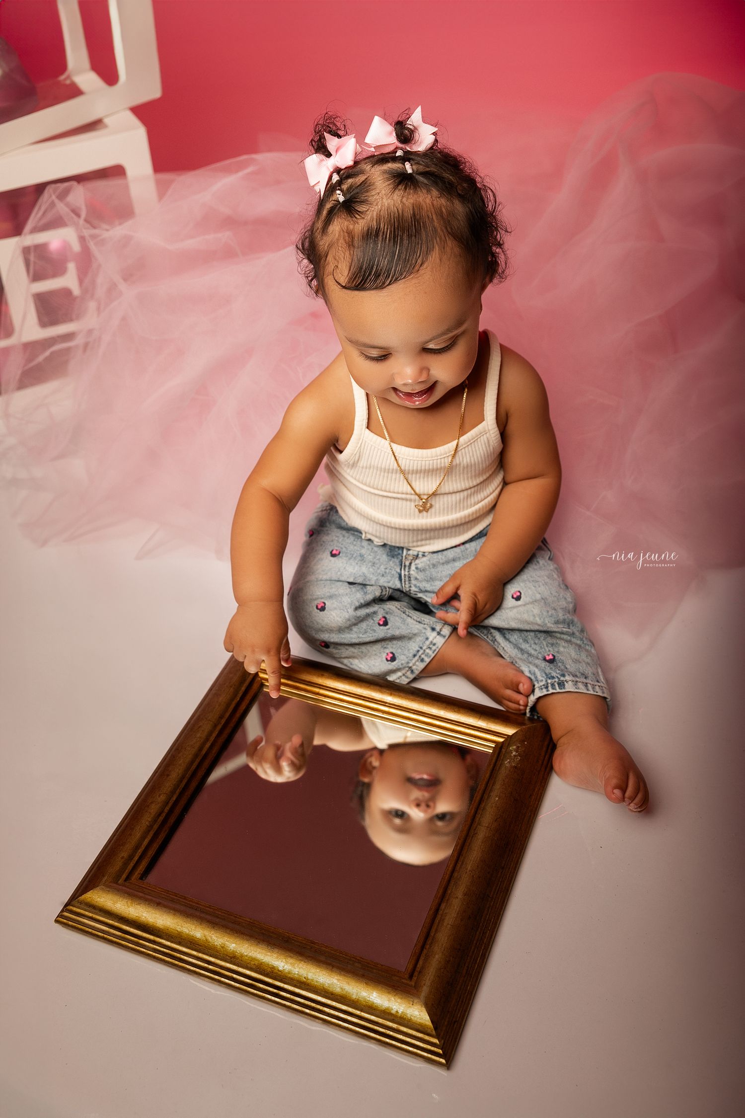Young child in casual attire plays with a golden picture frame against pink backdrop.