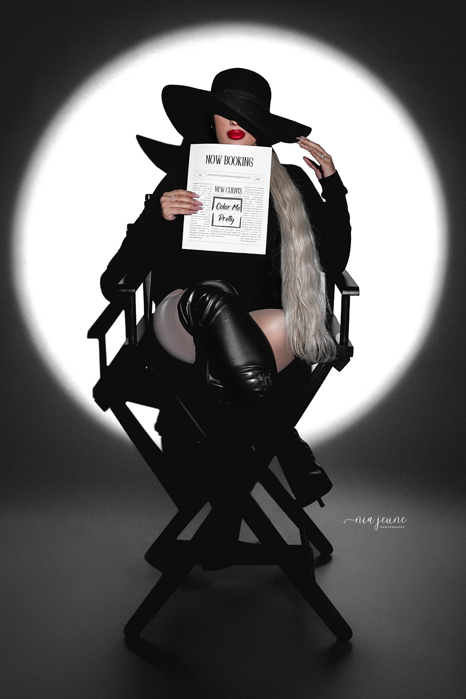 Stylish figure in black attire and wide-brimmed hat sits in director's chair holding script against dramatic white backlight.