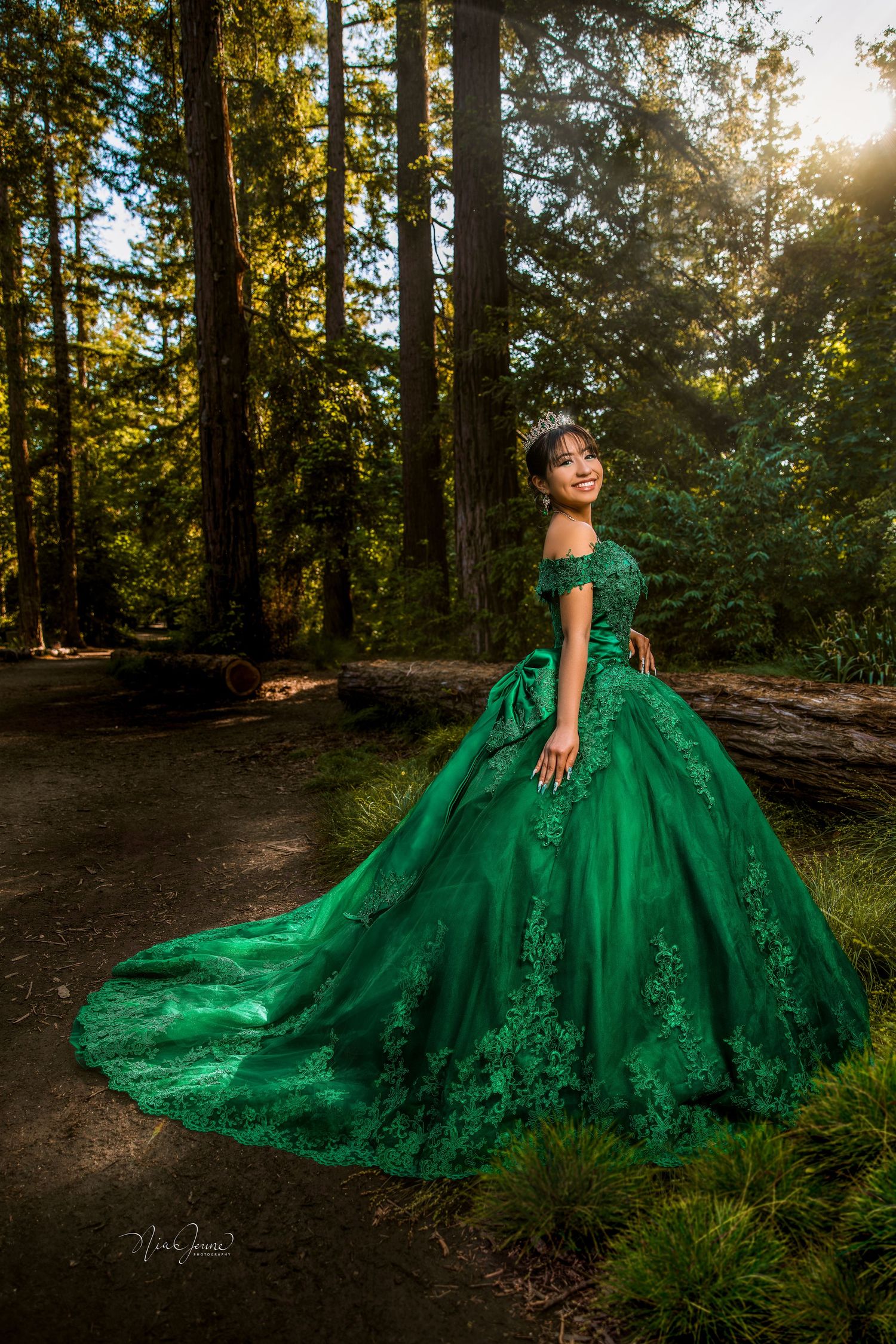 Elegant emerald green ball gown with off-shoulder design and floral details photographed in sunlit forest setting.