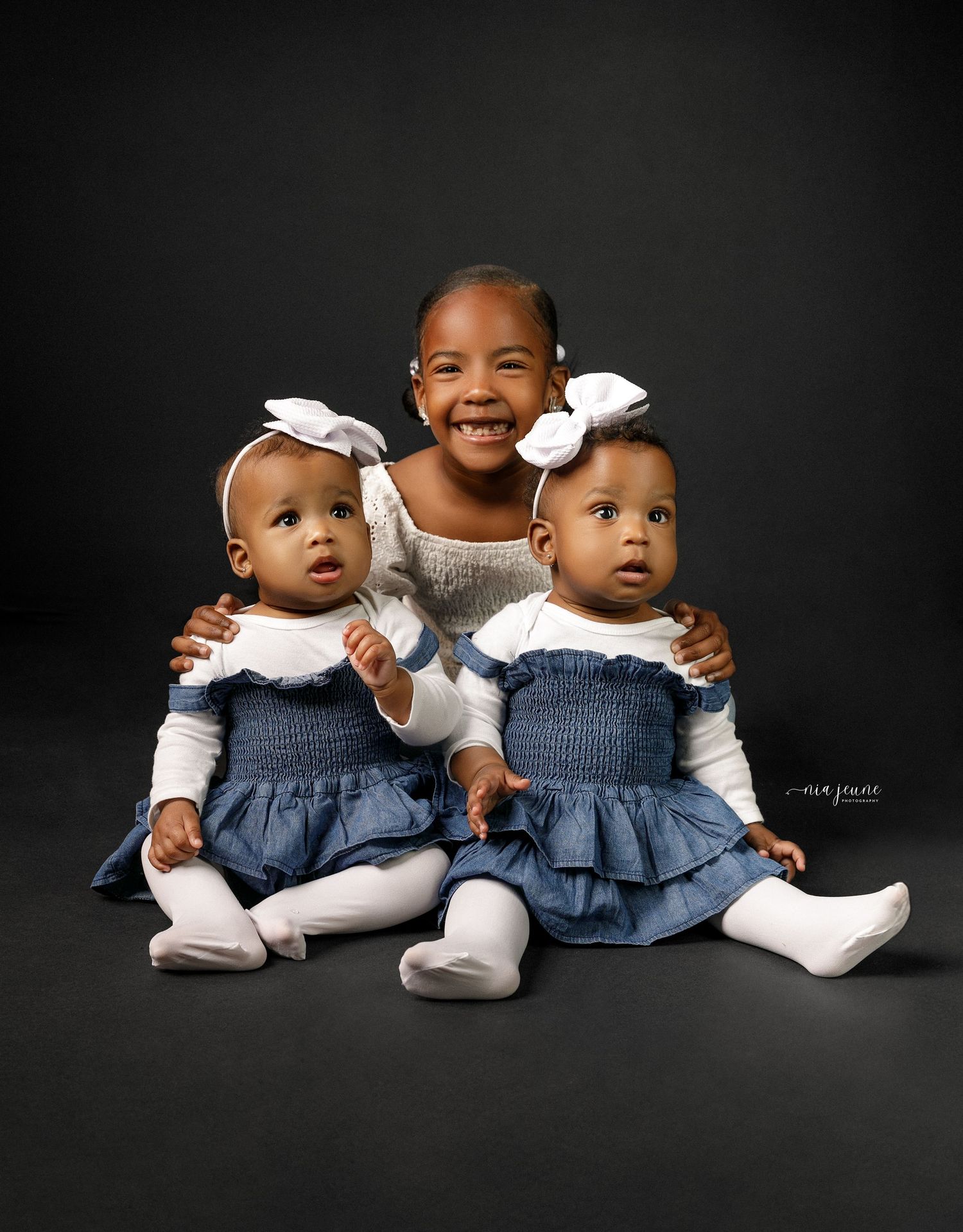 Studio portrait against black background showing three children in blue denim dresses and white tops huddled together.