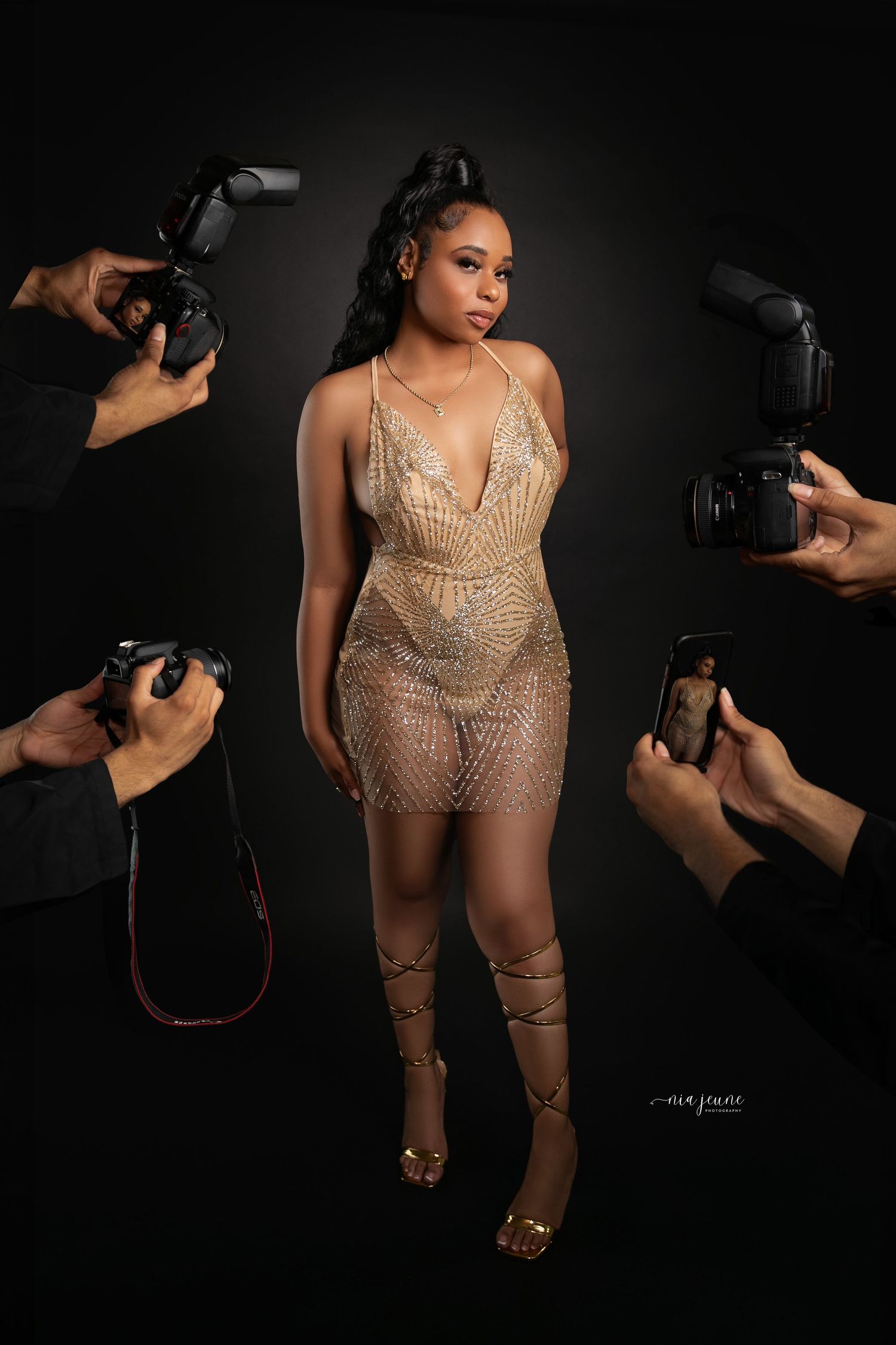 Behind-the-scenes shot of photographers capturing model in sheer gold dress against dark background.