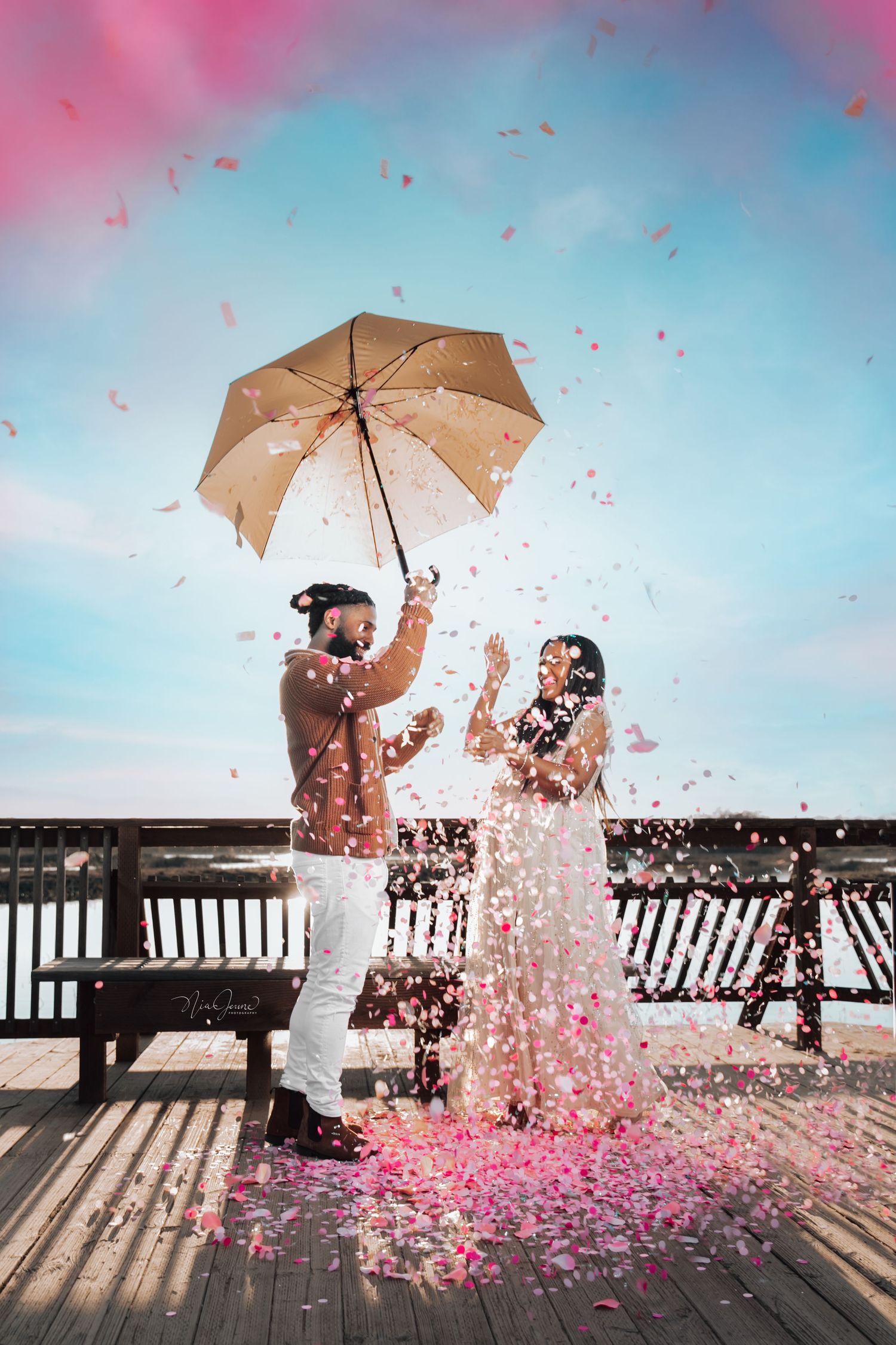 A couple celebrating on a wooden deck showered with pink flower petals while holding a beige umbrella against blue sky.