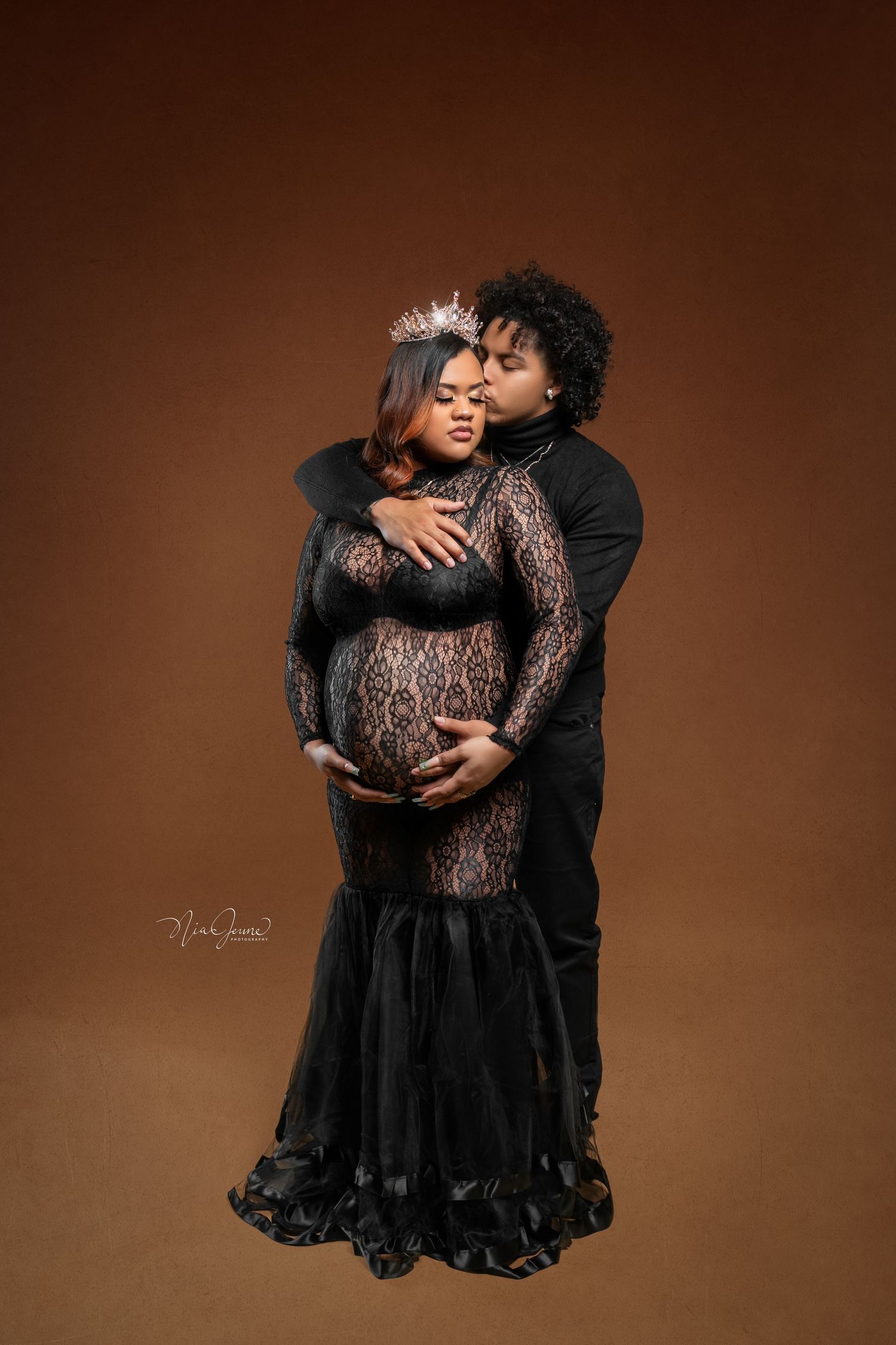 Romantic maternity portrait featuring couple in black formal attire against brown backdrop.