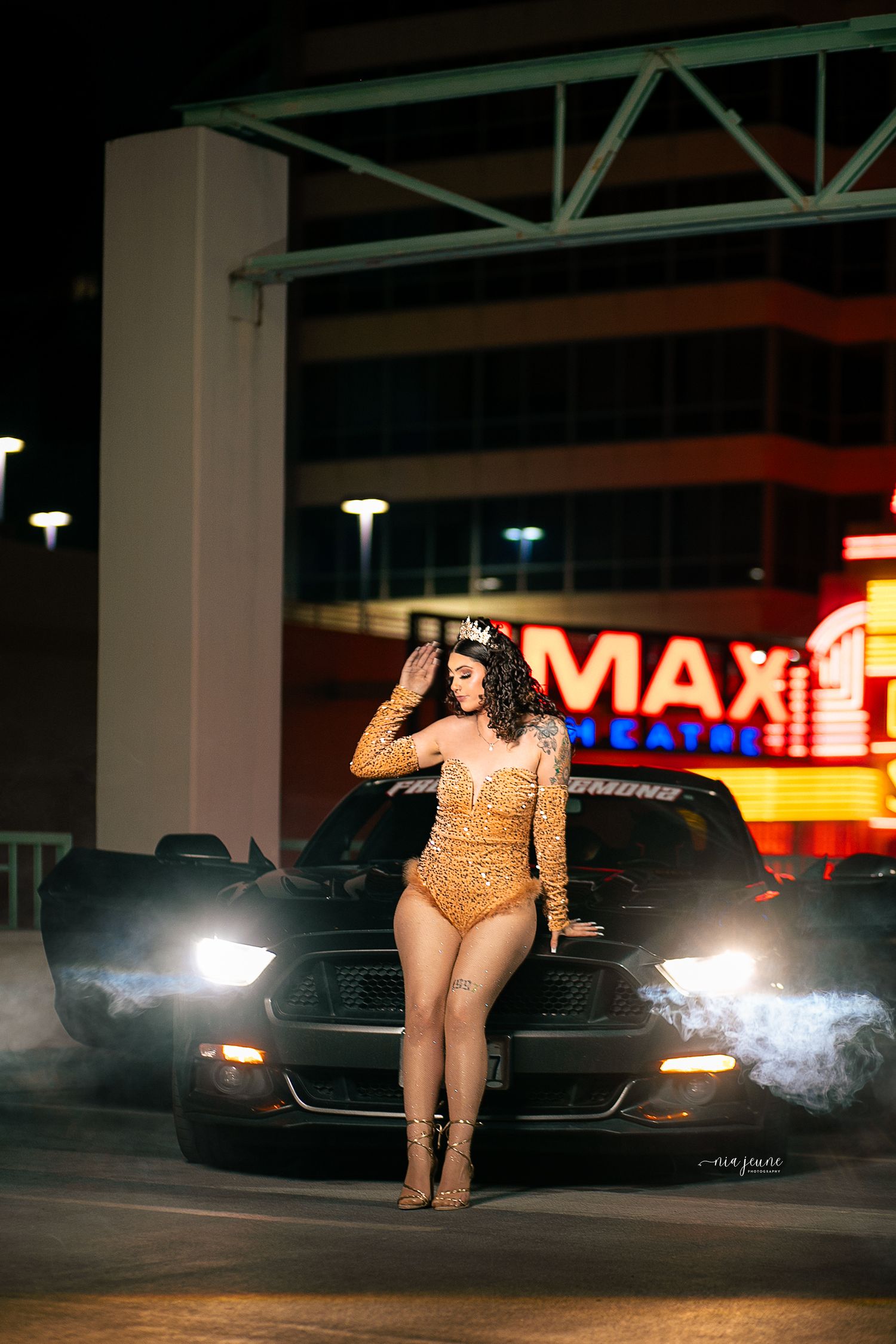 A person in a sparkly gold bodysuit poses in front of a black Mustang car with illuminated headlights at night.