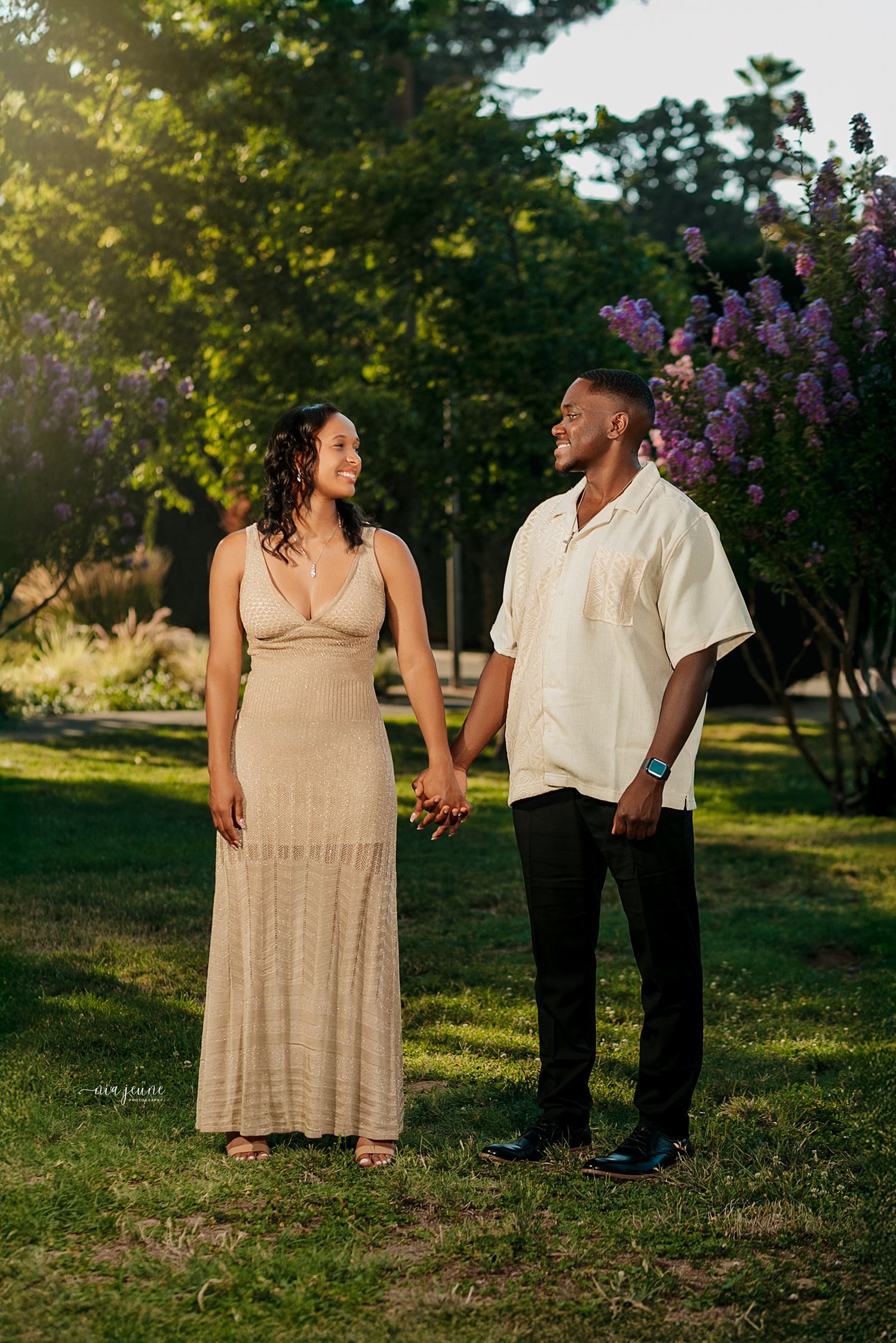 A couple holding hands and walking together in a garden setting during golden hour with flowering lilacs in the background.