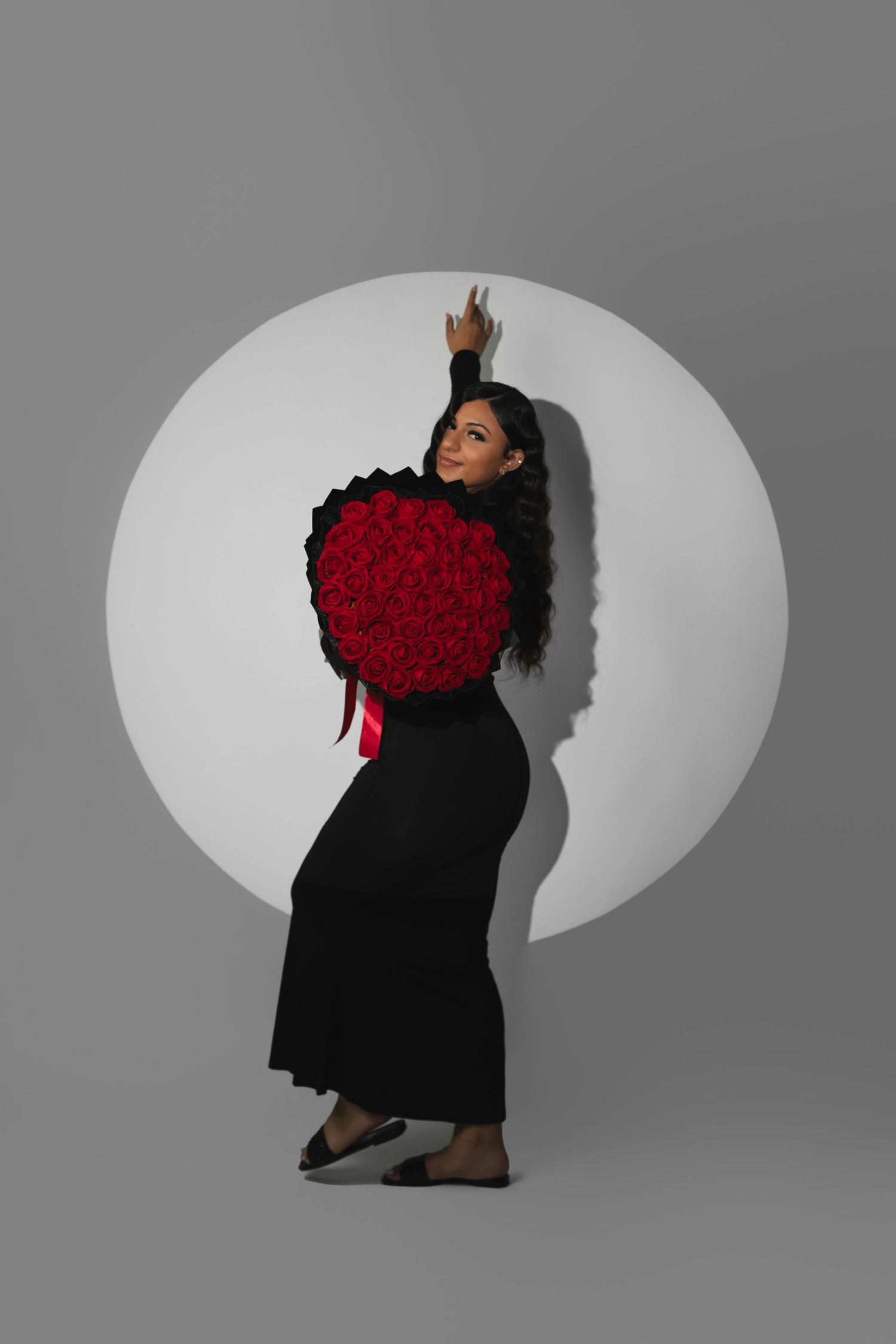 A figure in a flowing black dress poses dramatically with a bouquet of red roses against a light gray studio backdrop.
