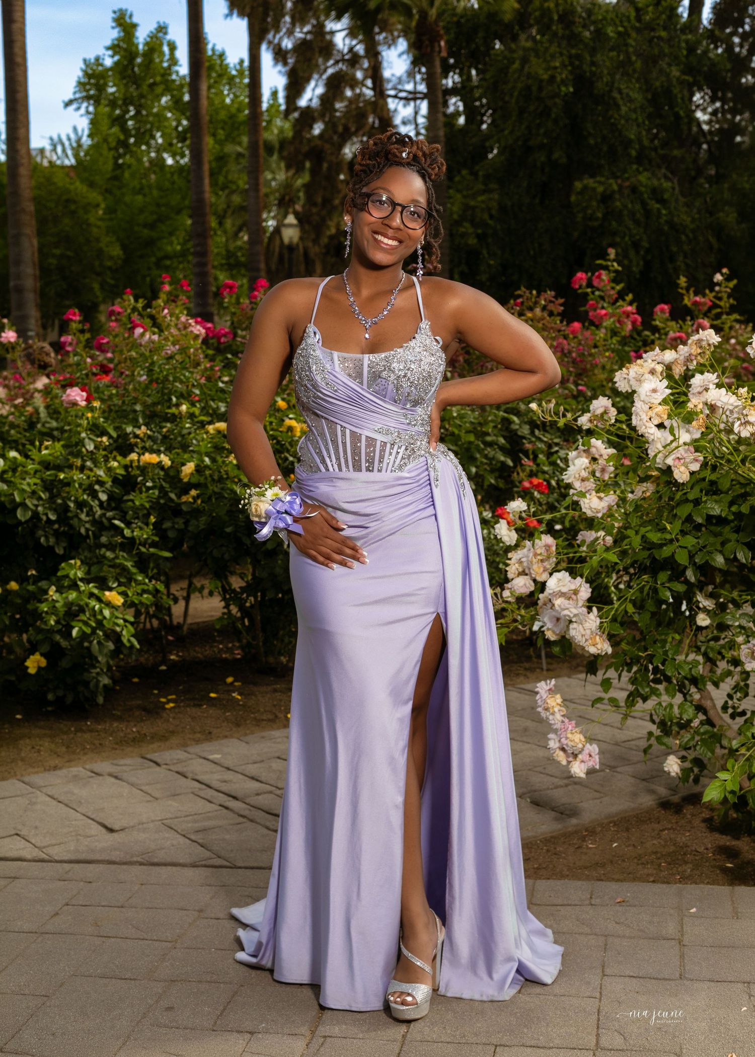 Young woman in lavender prom gown with crystal bodice poses among colorful roses in garden.