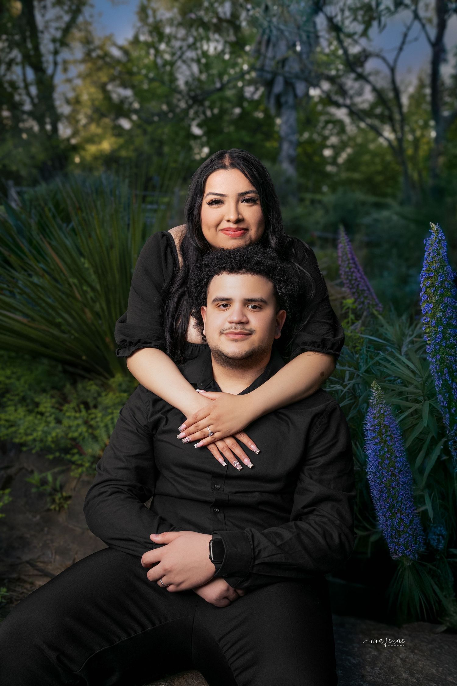 Portrait of a couple wearing black clothing embracing in a garden with purple flowers.