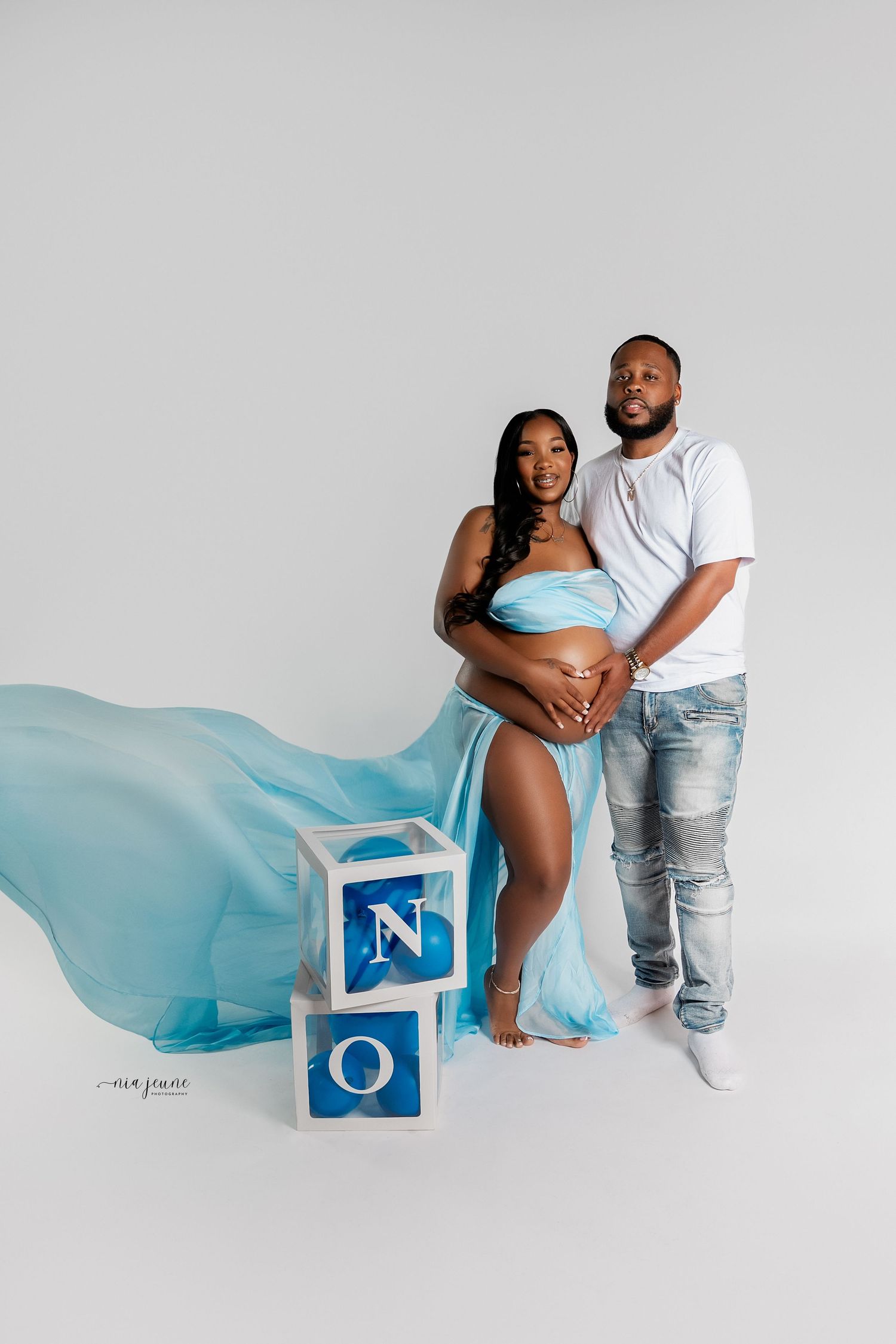 Maternity photo with flowing turquoise fabric and decorative baby blocks on white background.