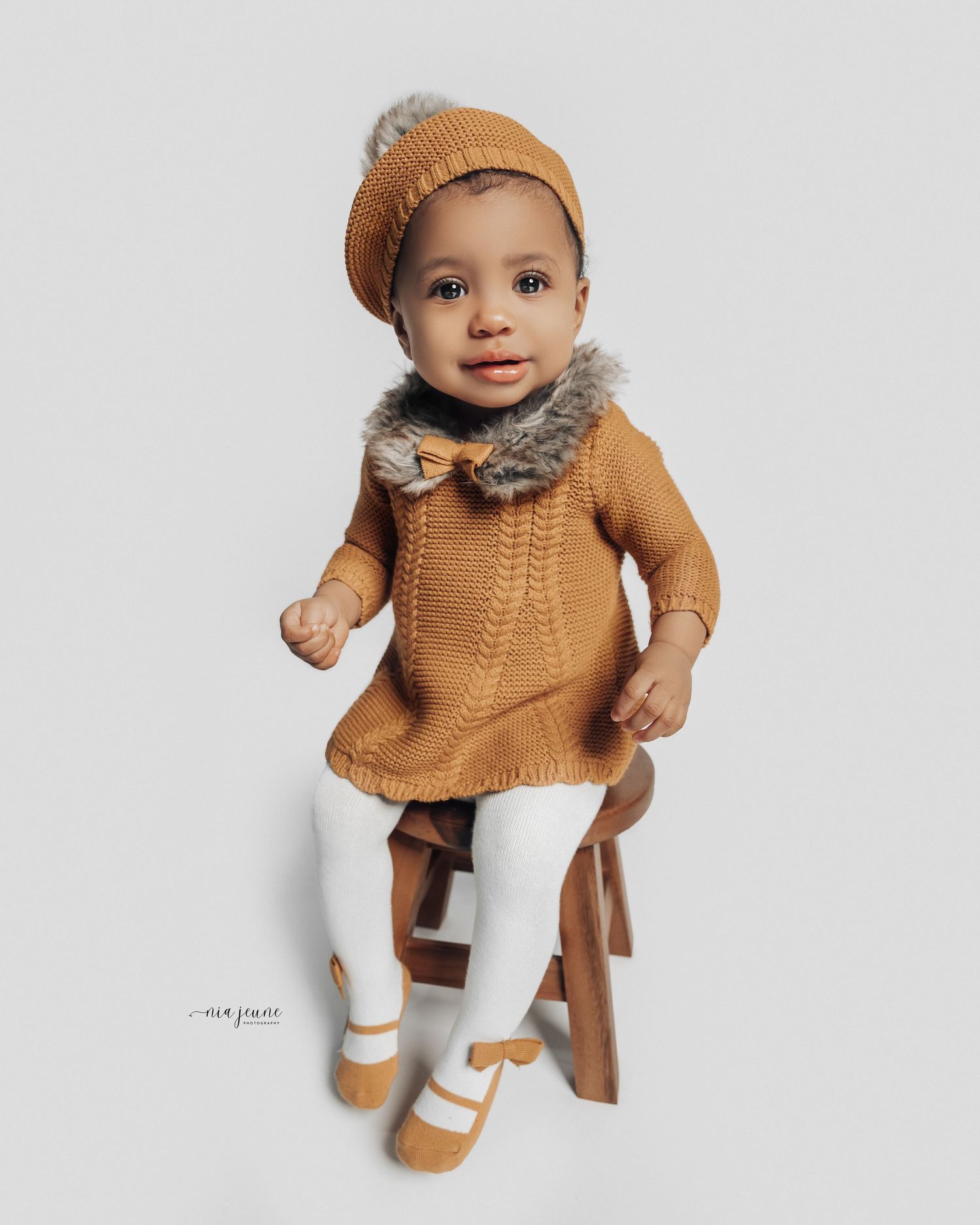 Baby doll wearing cozy brown knit sweater and white pants sits on wooden chair.