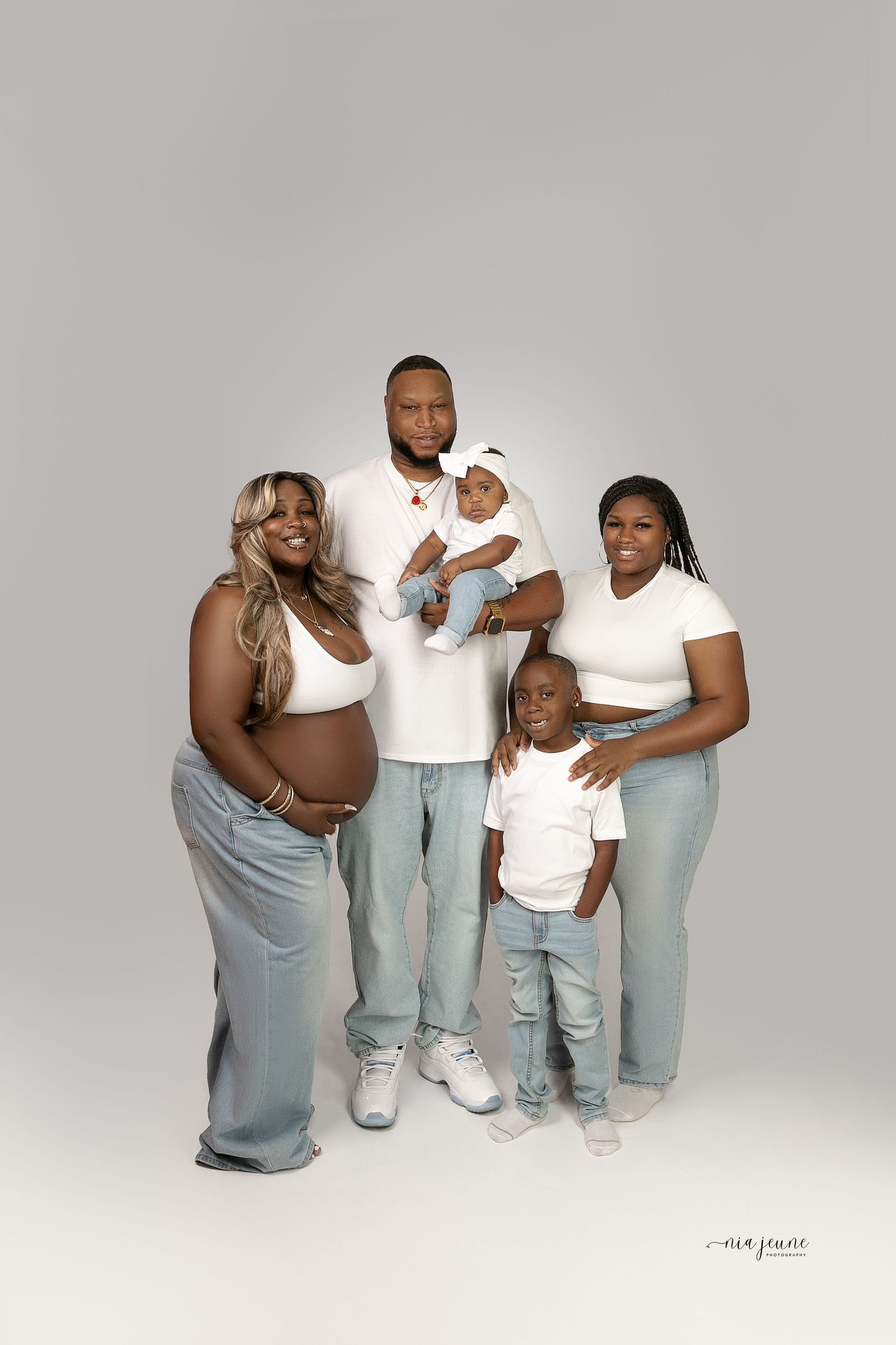 A loving family portrait in coordinated white and denim outfits against a grey studio background.