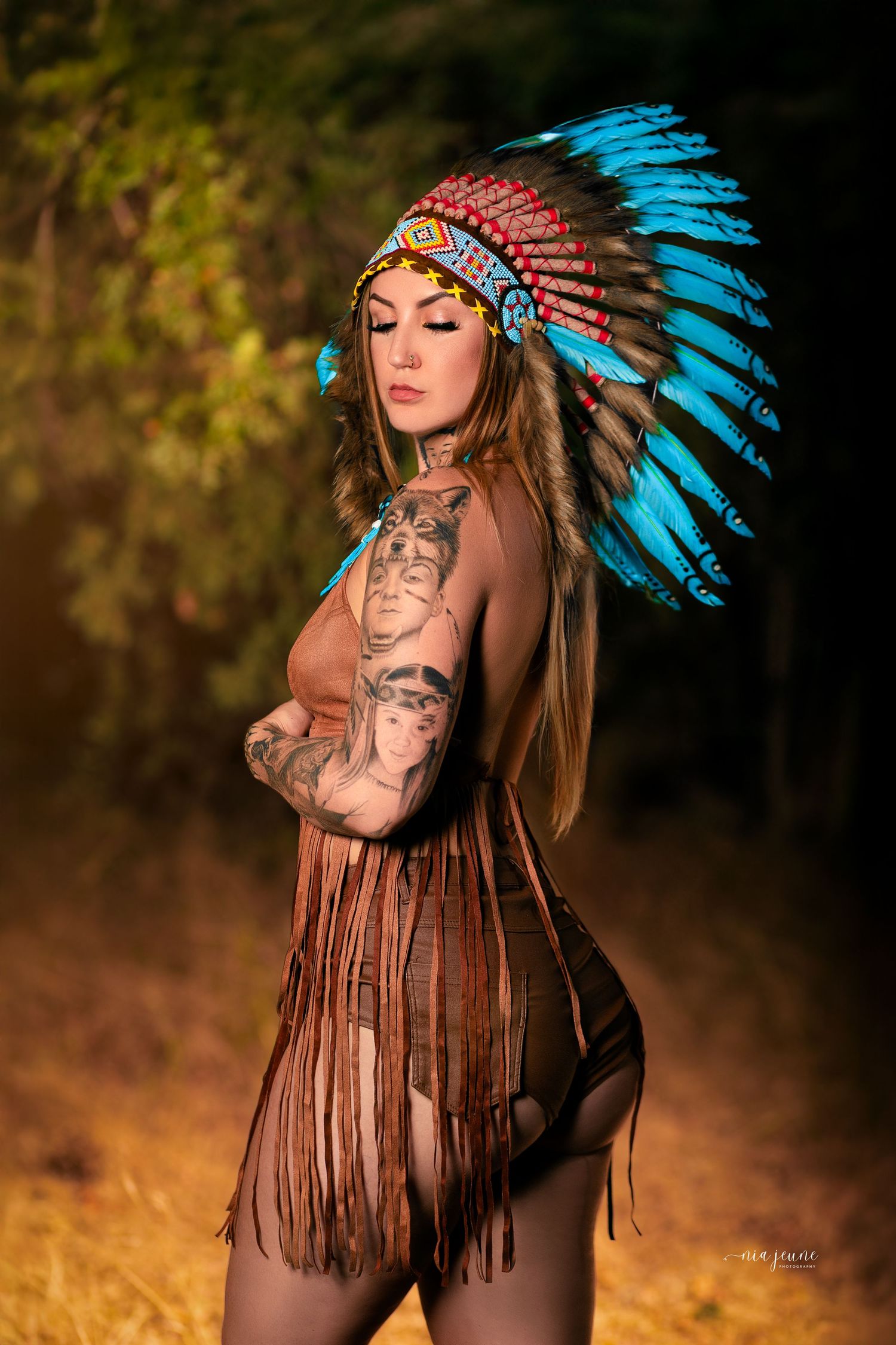 A model wearing a turquoise and red Native American headdress and fringed leather dress poses in golden sunlight.