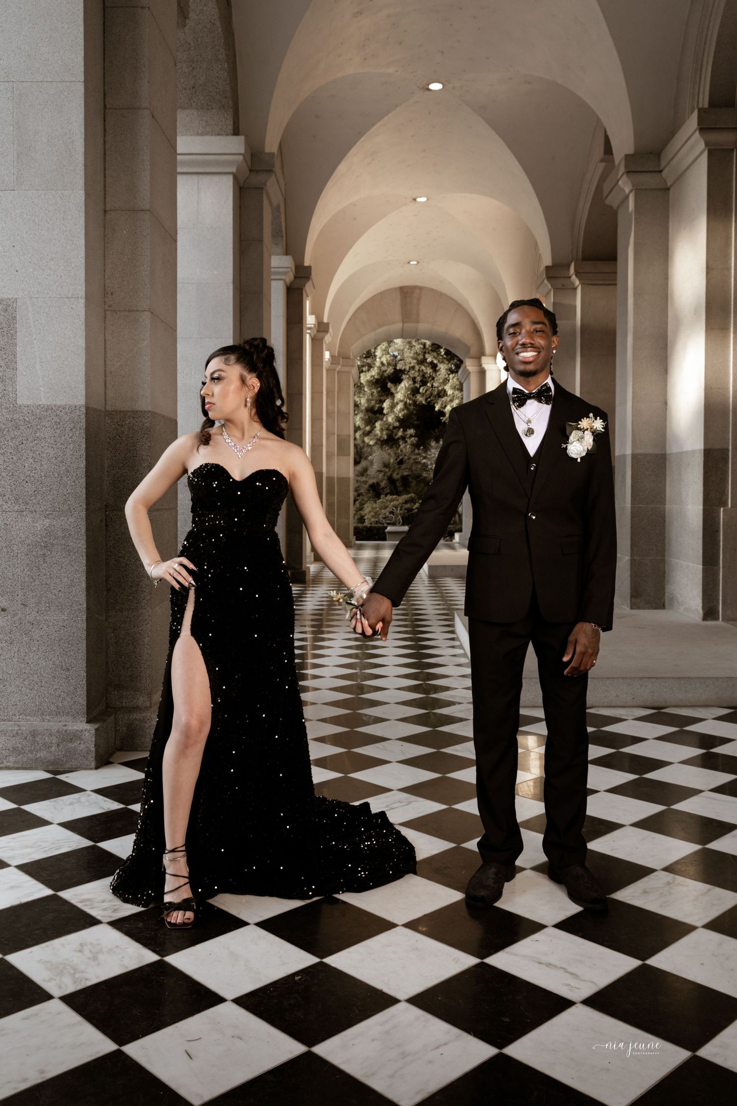 Elegant couple in formal wear posing on checkered floor beneath arched hallway.