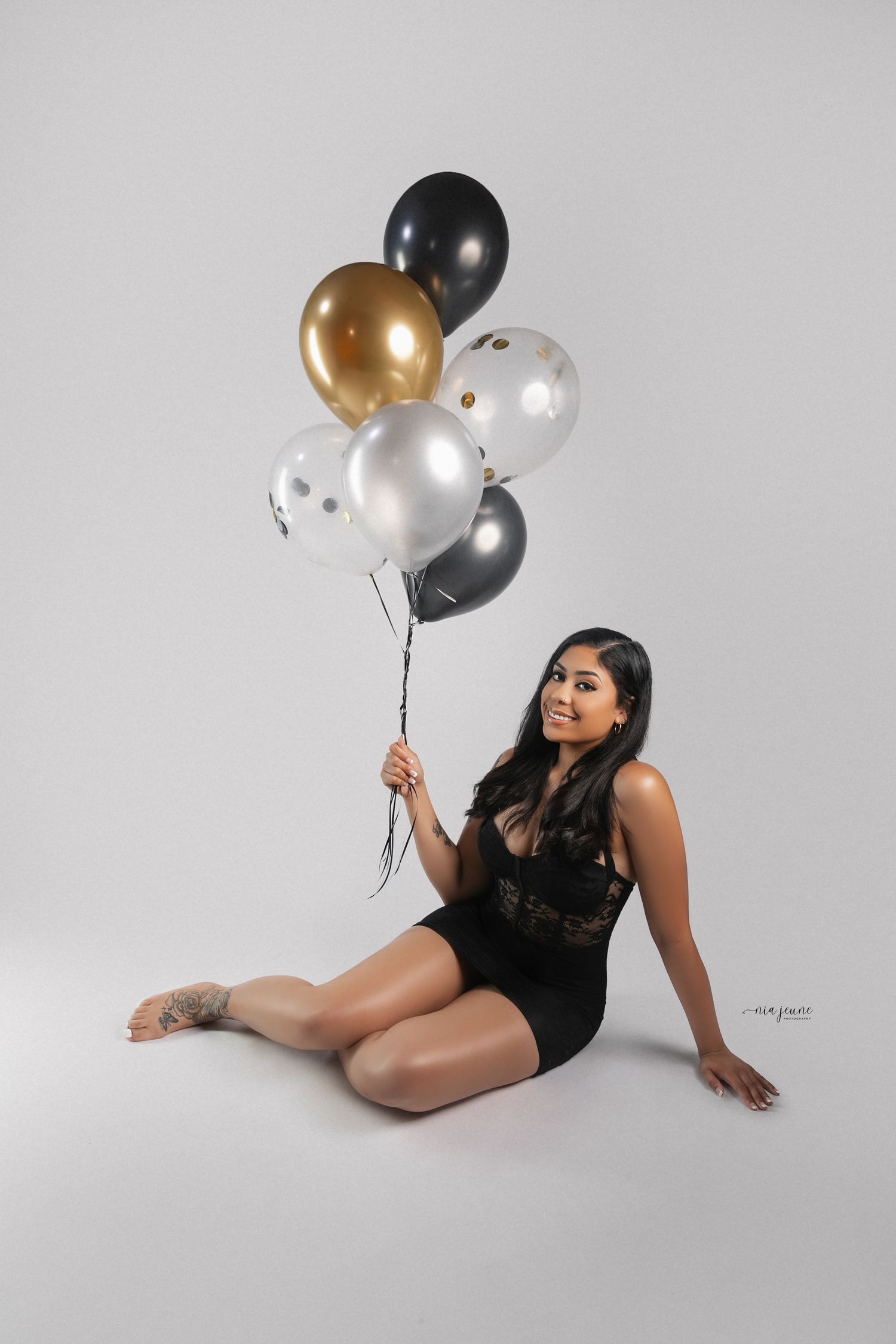 Model in black bodysuit poses elegantly with silver and gold balloons against white background.