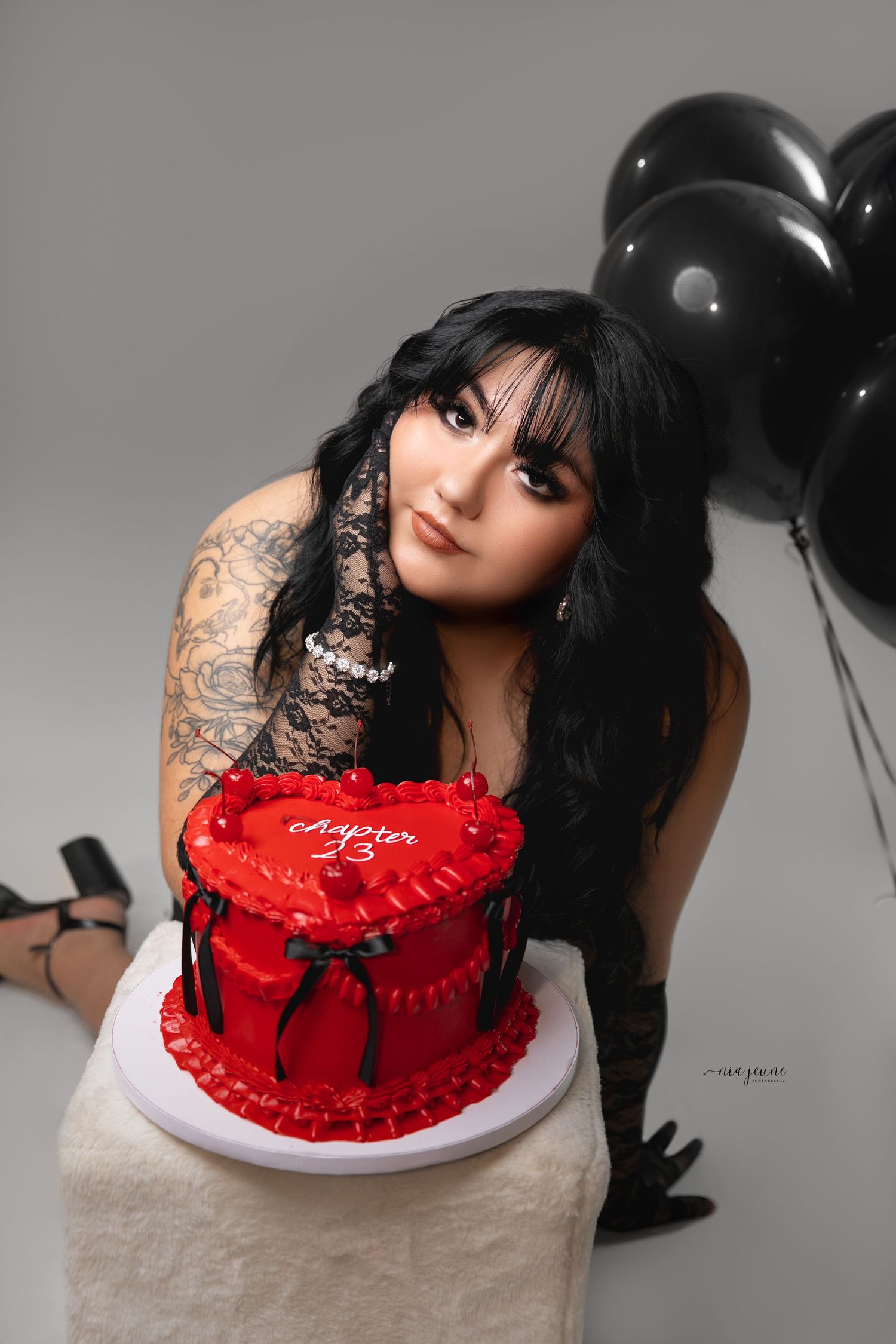 Moody portrait with black lace, red cake, and black balloons in grayscale setting.
