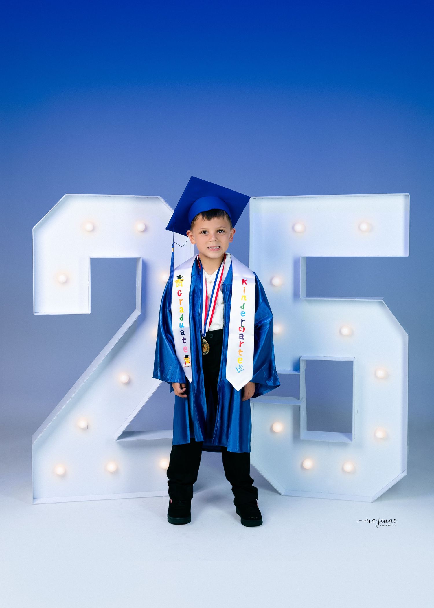 Young graduate in blue cap and gown stands proudly in front of illuminated '25' display.