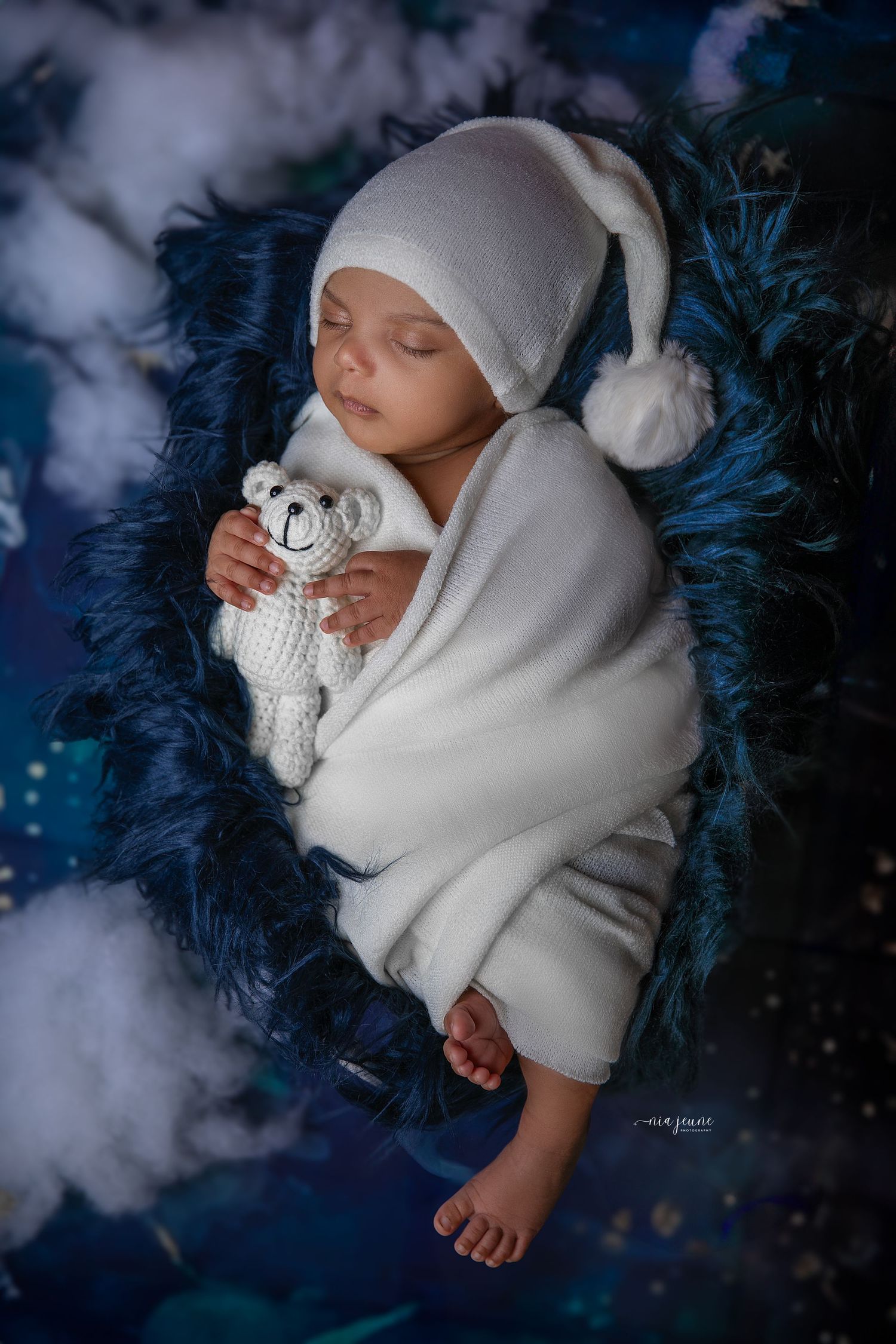 Sleeping baby wrapped in white winter clothing holding a teddy bear against a magical blue background.