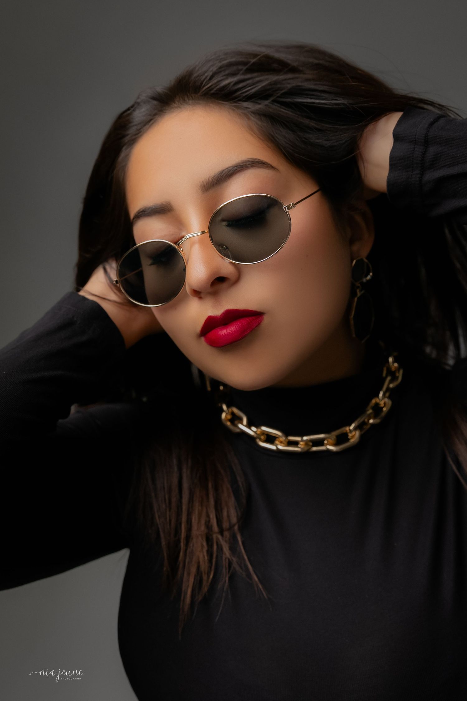 Glamorous portrait with aviator sunglasses, red lipstick, and gold chain necklace against dark background.