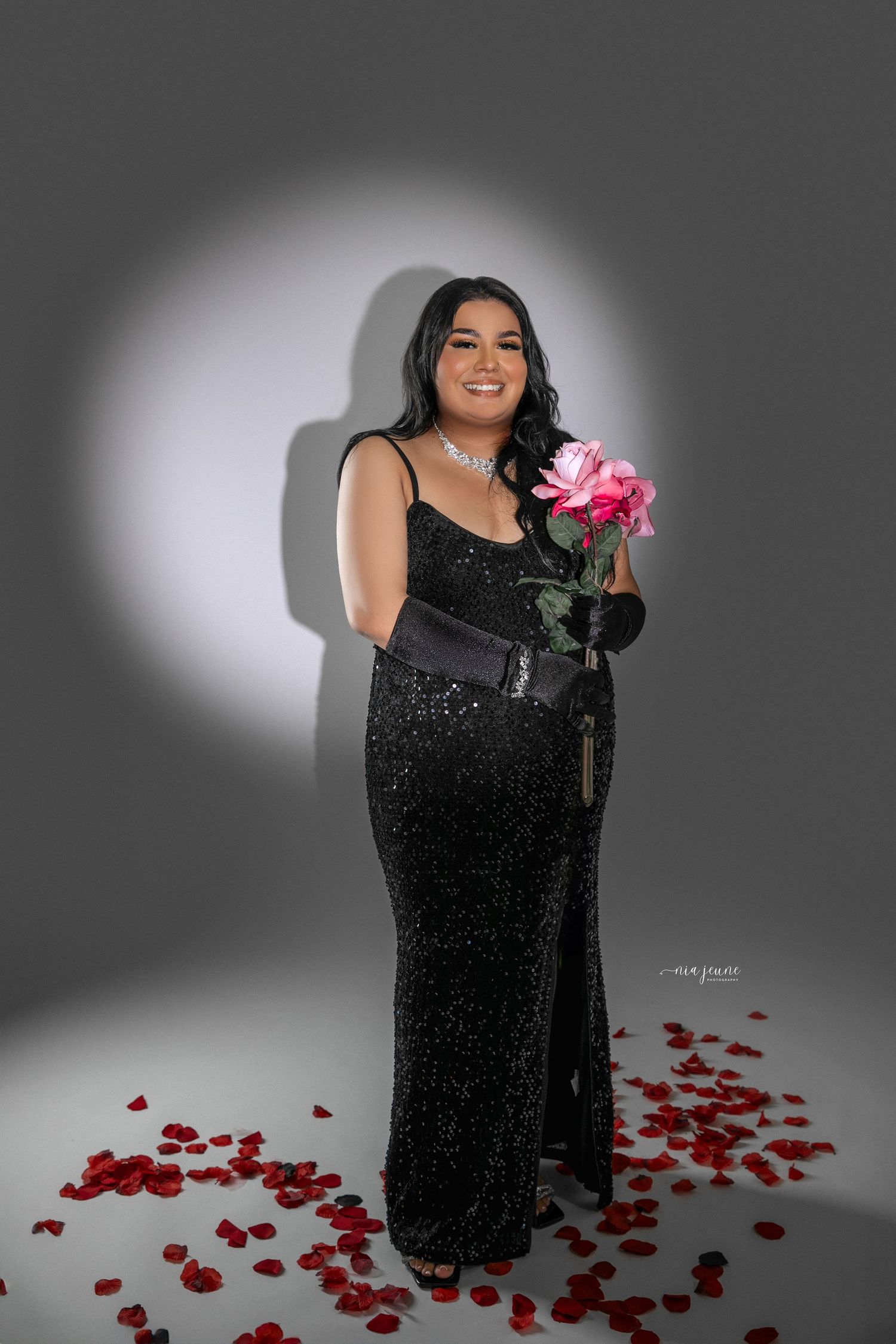 Model in sparkly black evening gown holds pink rose while standing on rose petal covered floor.