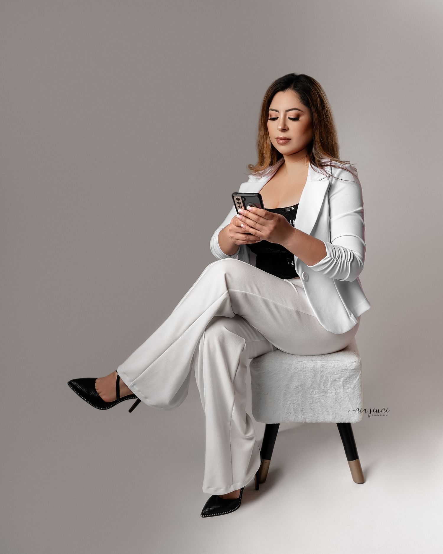 Professional woman in white business suit sitting elegantly in chair while checking phone.