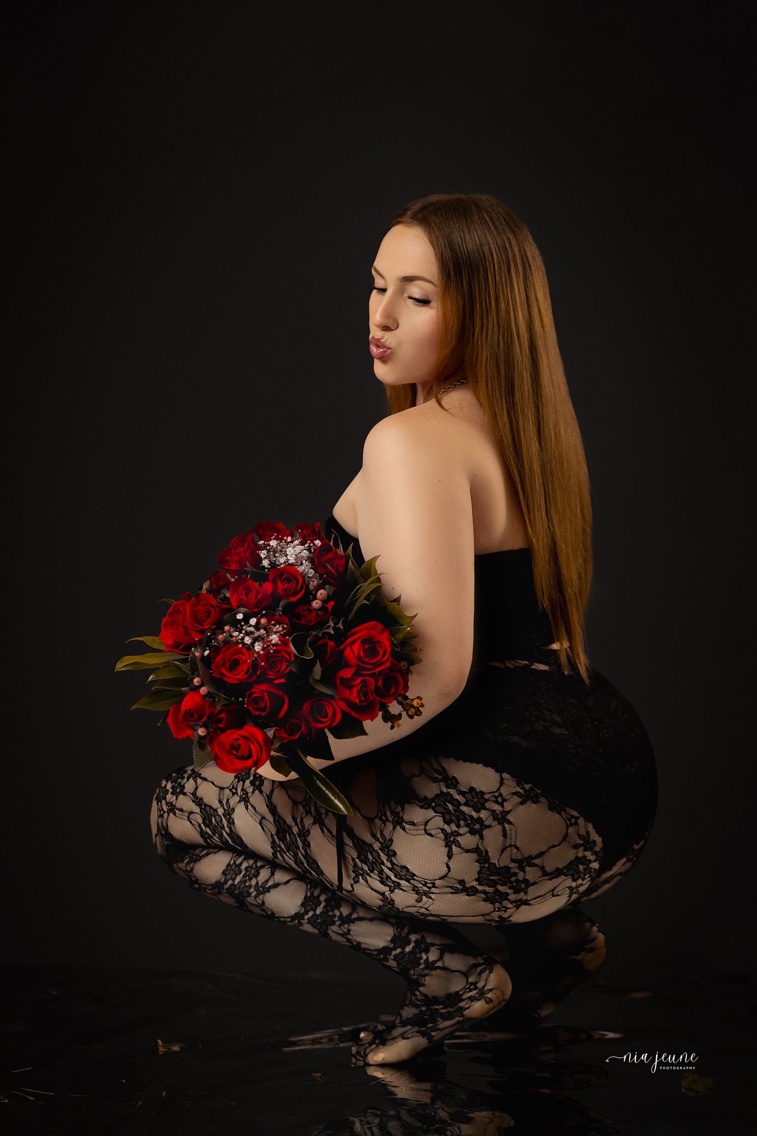 A figure in black lace holds a bouquet of deep red roses against a dark background in a sensual boudoir photoshoot.