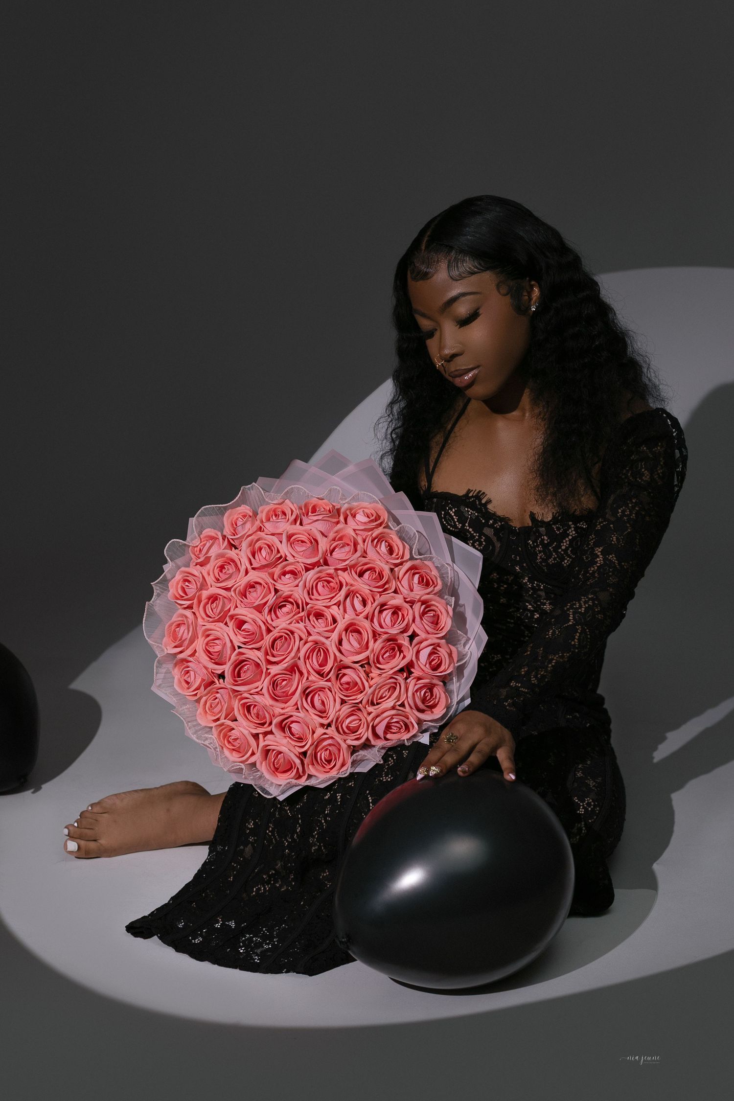 Woman in black lace dress holding large pink rose bouquet with black balloon, studio portrait.