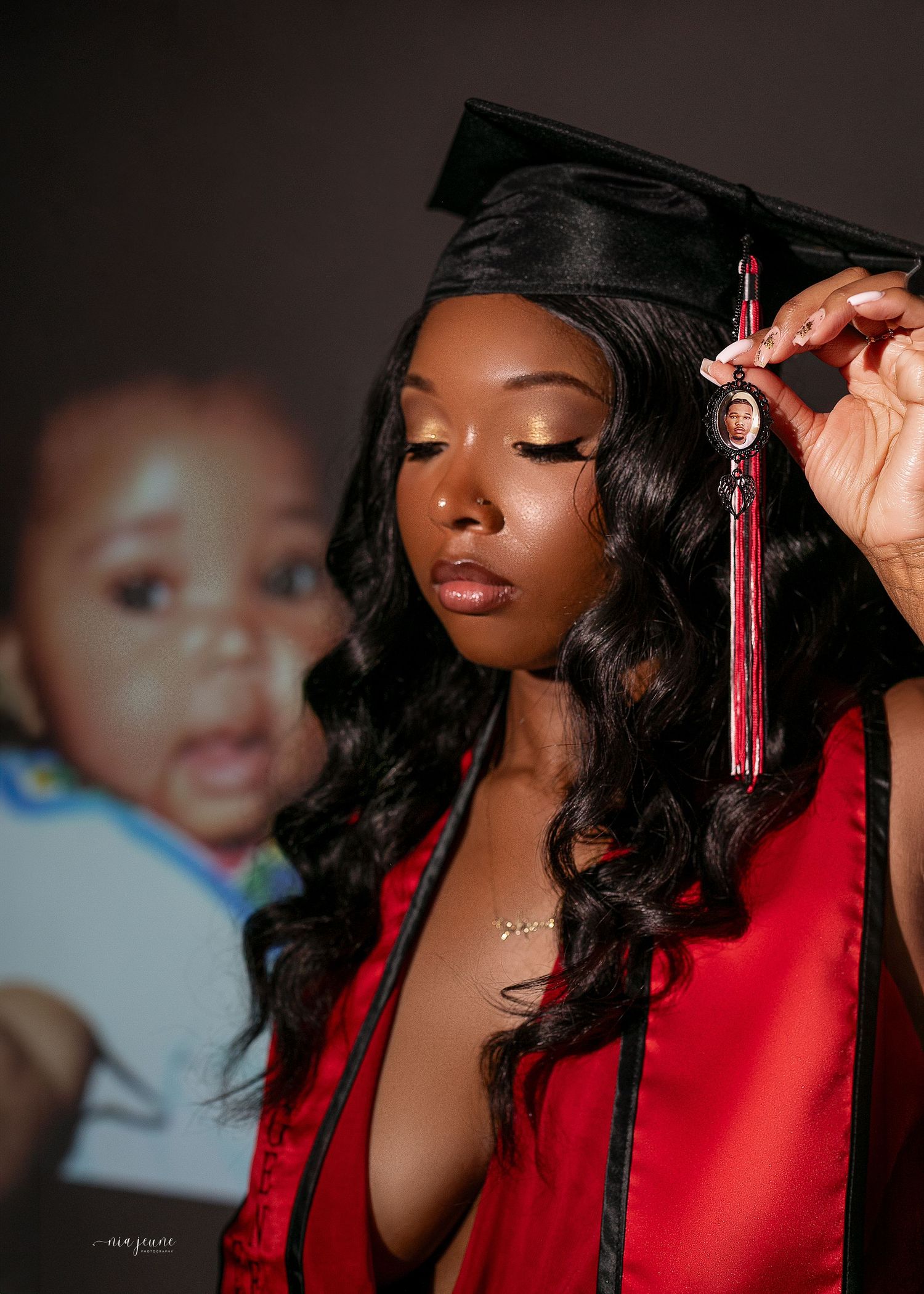 Young woman in red graduation cap and gown holding tassel with baby photo charm, with infant portrait in background.