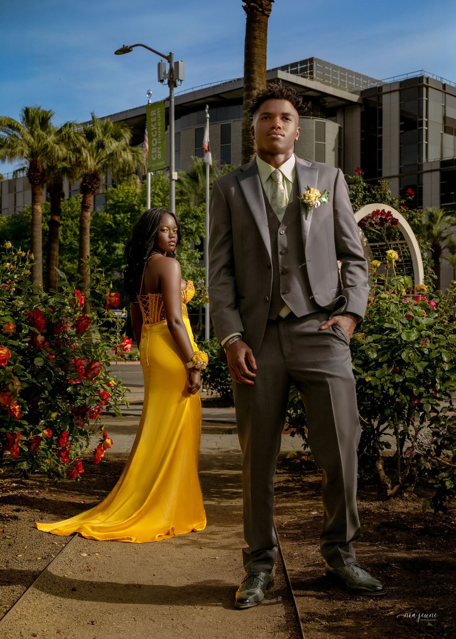 Couple in formal wear posing outdoors, woman in yellow gown and man in gray suit with boutonniere among roses.