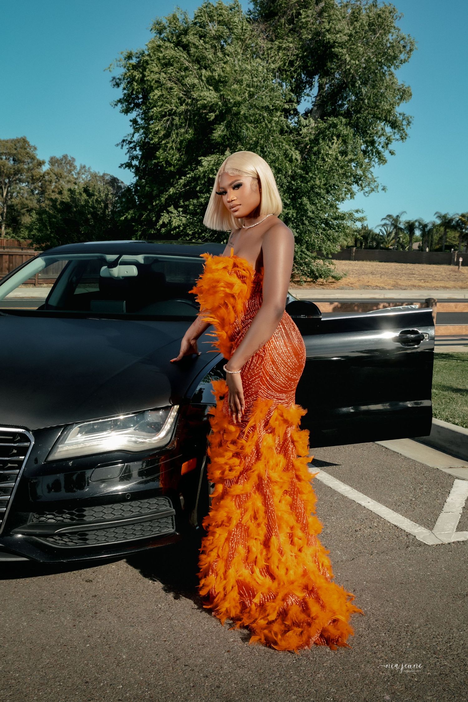 Person in stunning orange feathered gown poses next to black luxury car.
