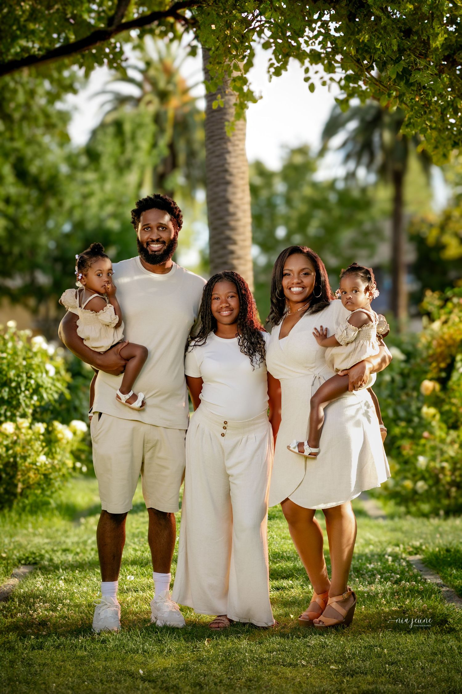 Family wearing coordinated white outfits poses together in a lush green outdoor setting.