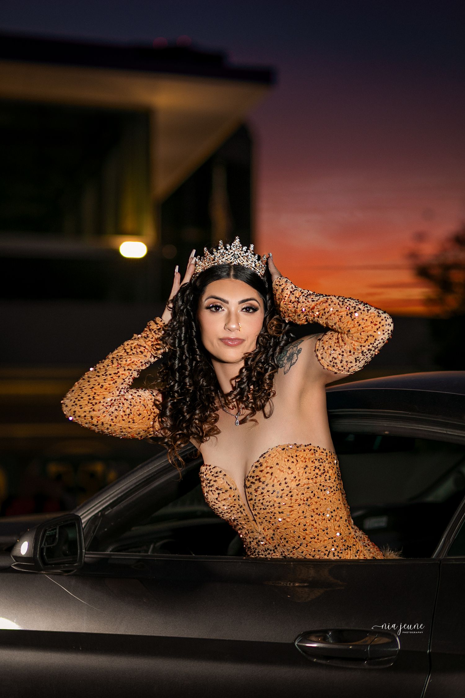 A glamorous figure in a sparkling gold dress poses with a car during sunset creating dramatic evening portraits.