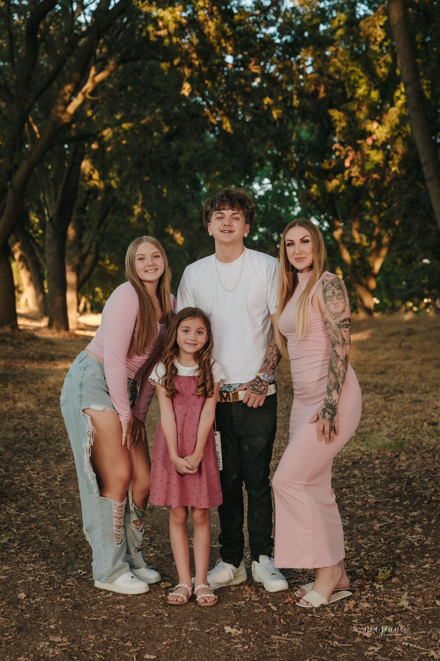 A group of siblings in coordinated pink and white outfits pose together for photos in a sunlit wooded area at sunset.