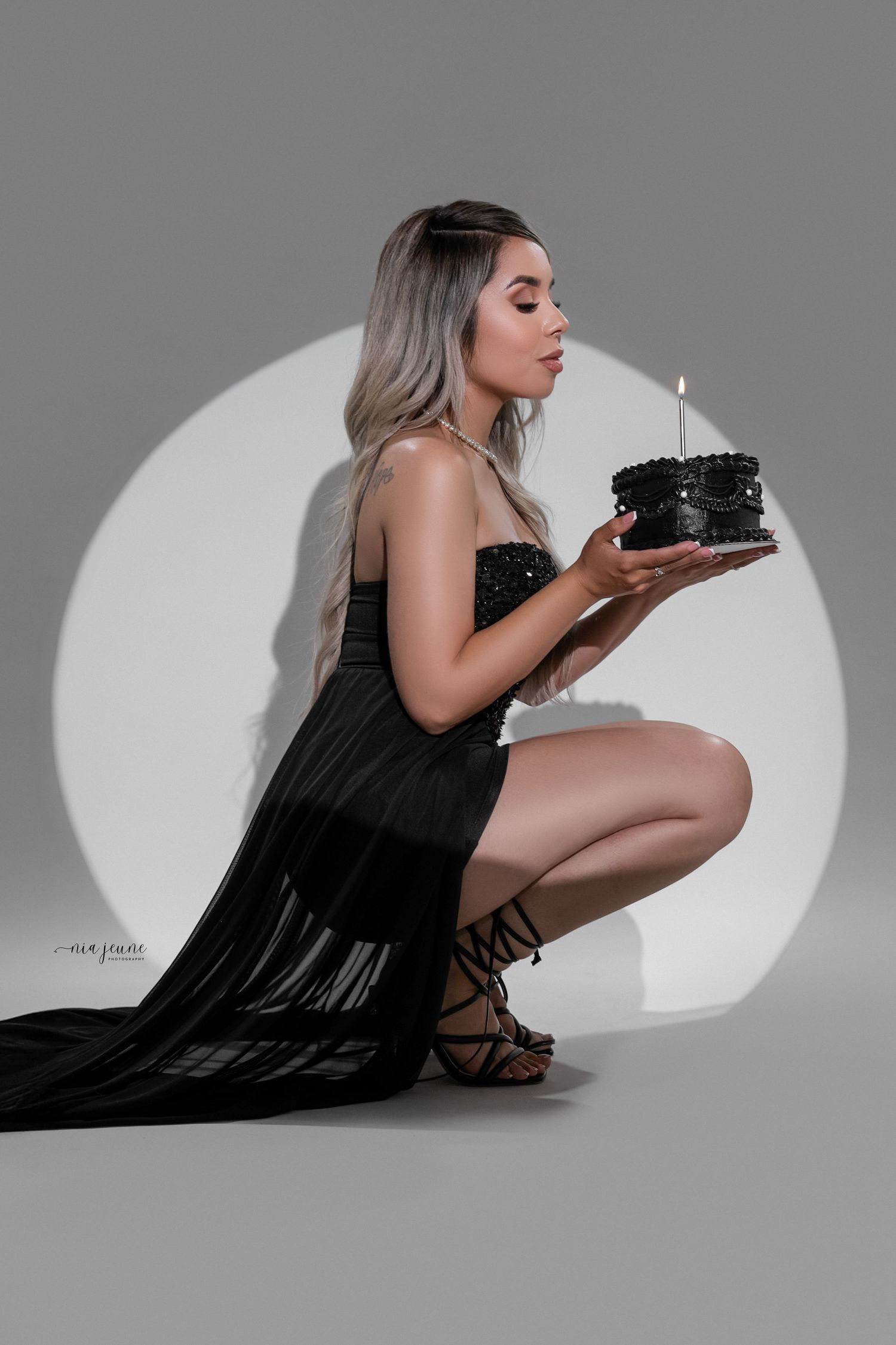 Dramatic portrait of model in black gown blowing out candle on dark cake against grey backdrop.