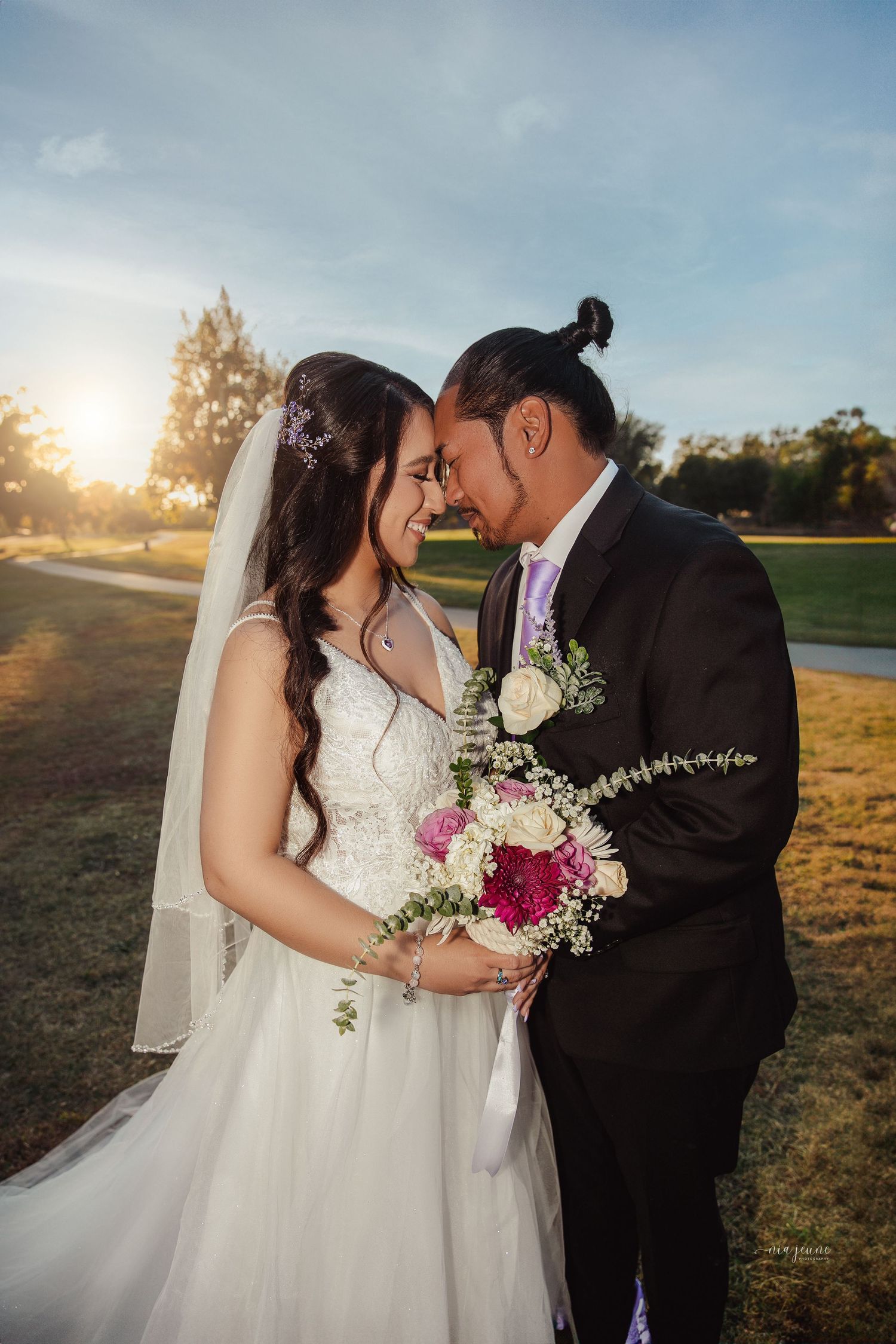 A romantic wedding photo of a couple sharing an intimate moment at sunset with a pink floral bouquet.