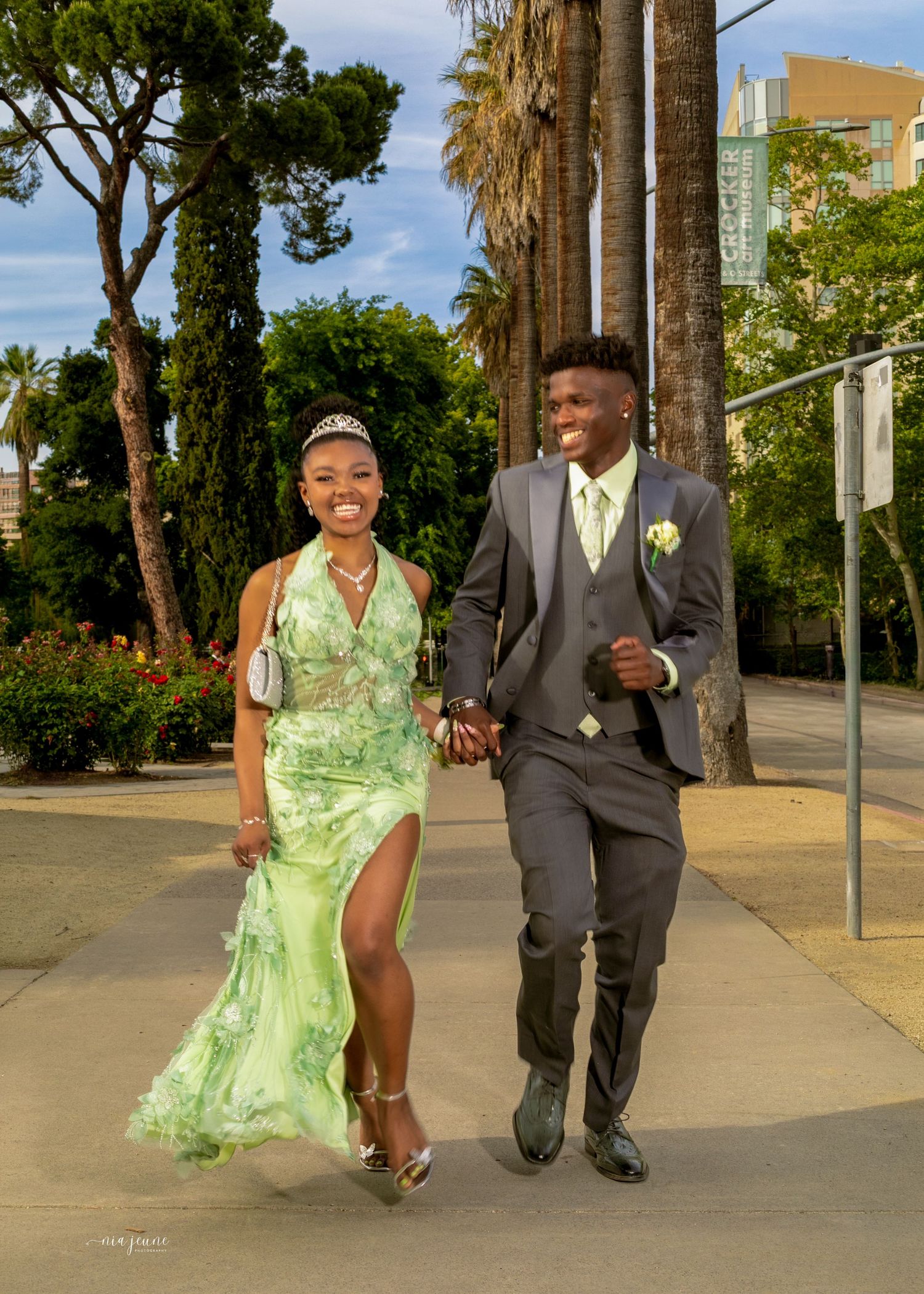 Elegant couple in matching green floral dress and gray suit walking hand-in-hand on a palm-lined sidewalk at prom.