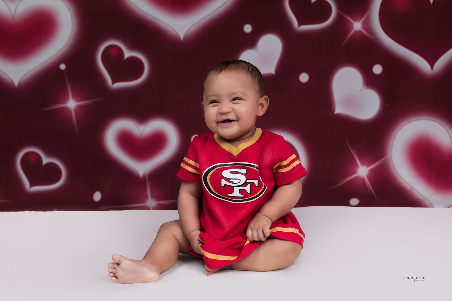 Smiling baby in a San Francisco 49ers red onesie sits against a Valentine's Day heart backdrop.