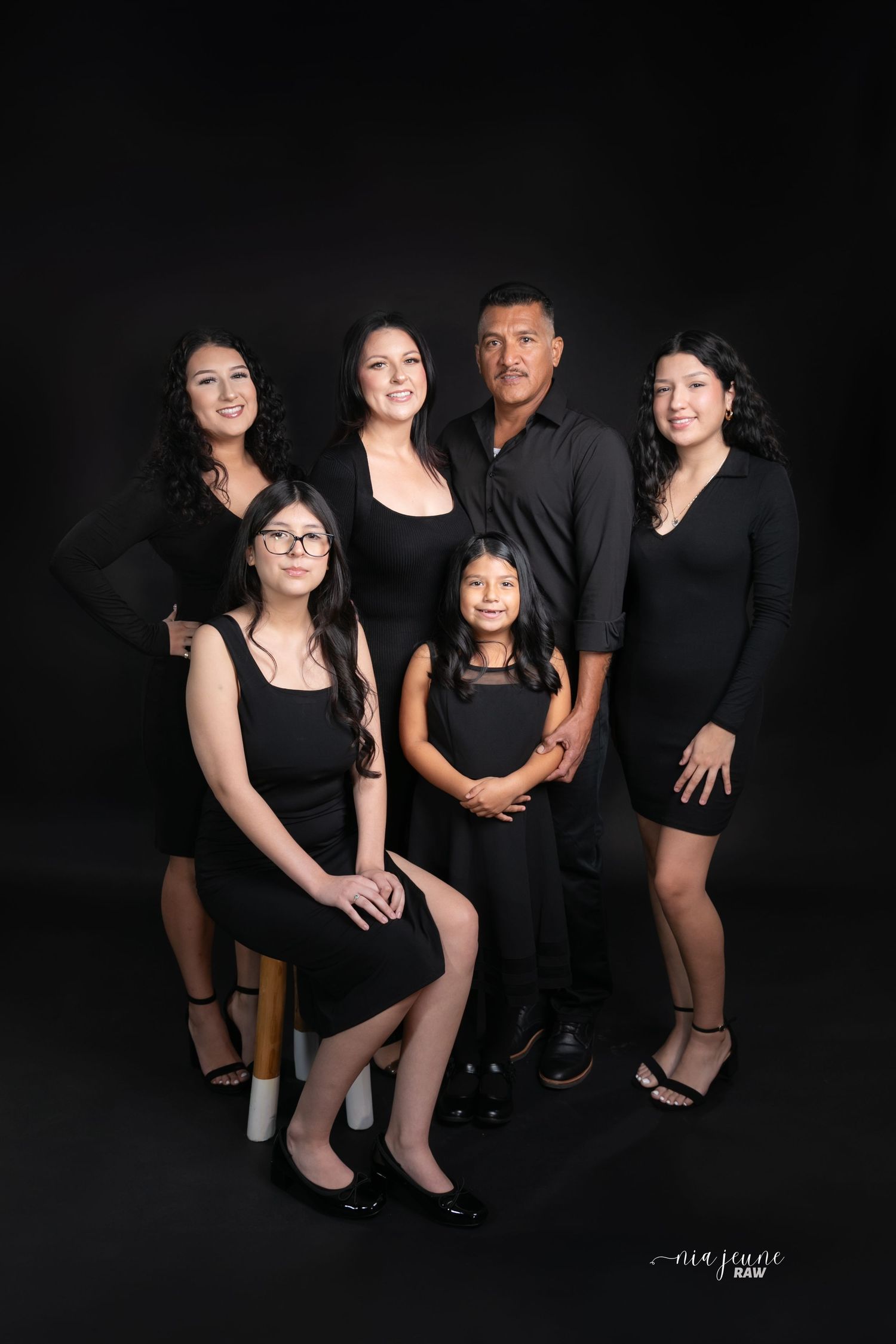 A formal family portrait of six people wearing black attire against dark background.