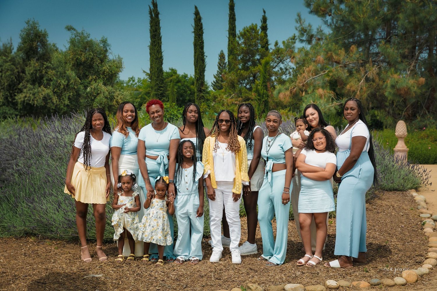 A group poses outdoors in coordinated white and light blue dresses against natural greenery and hillside backdrops.